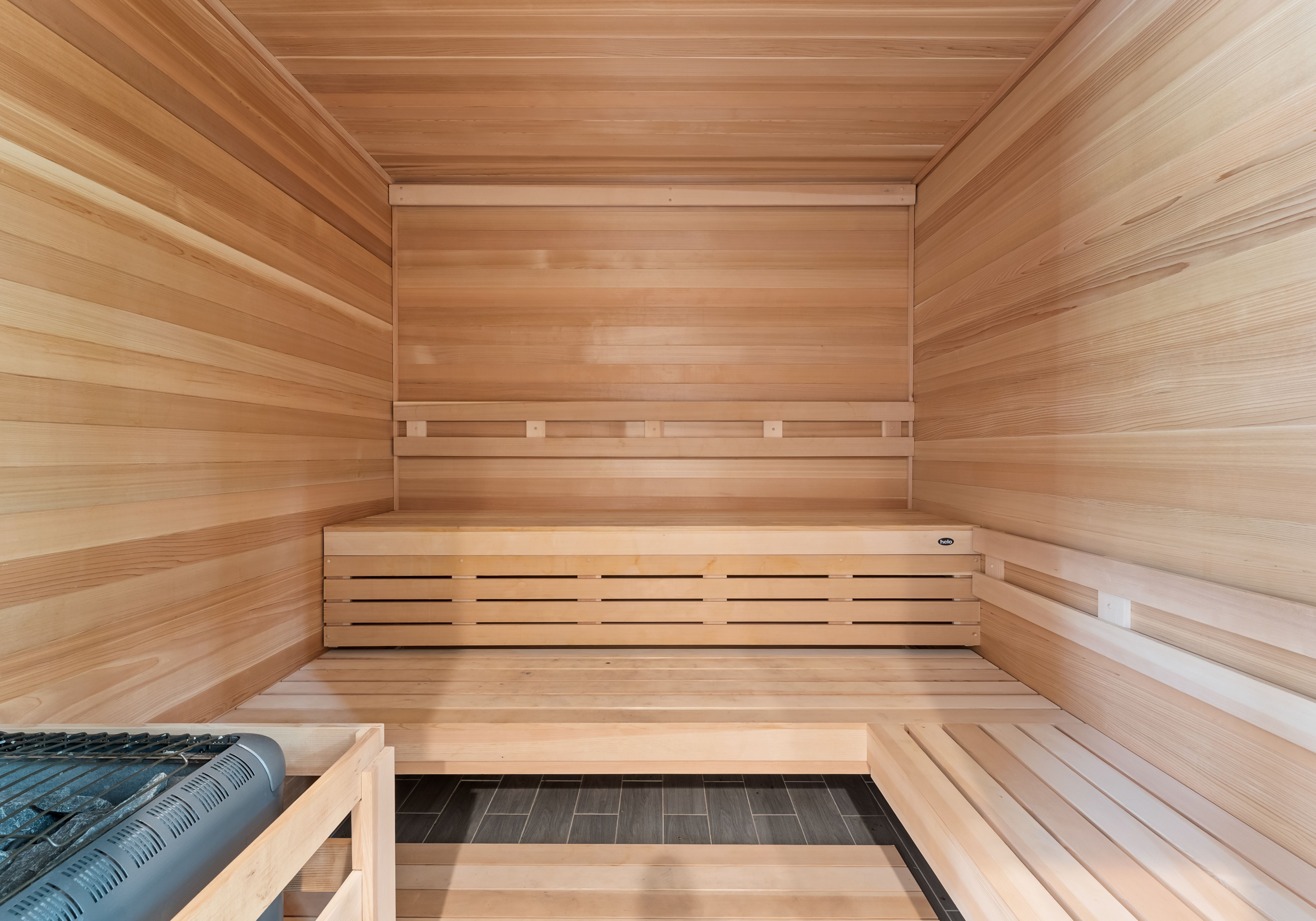 Interior of a sauna with light wooden walls and benches forming an L shape. A metal heater with stones sits in the bottom left corner on dark tile flooring.