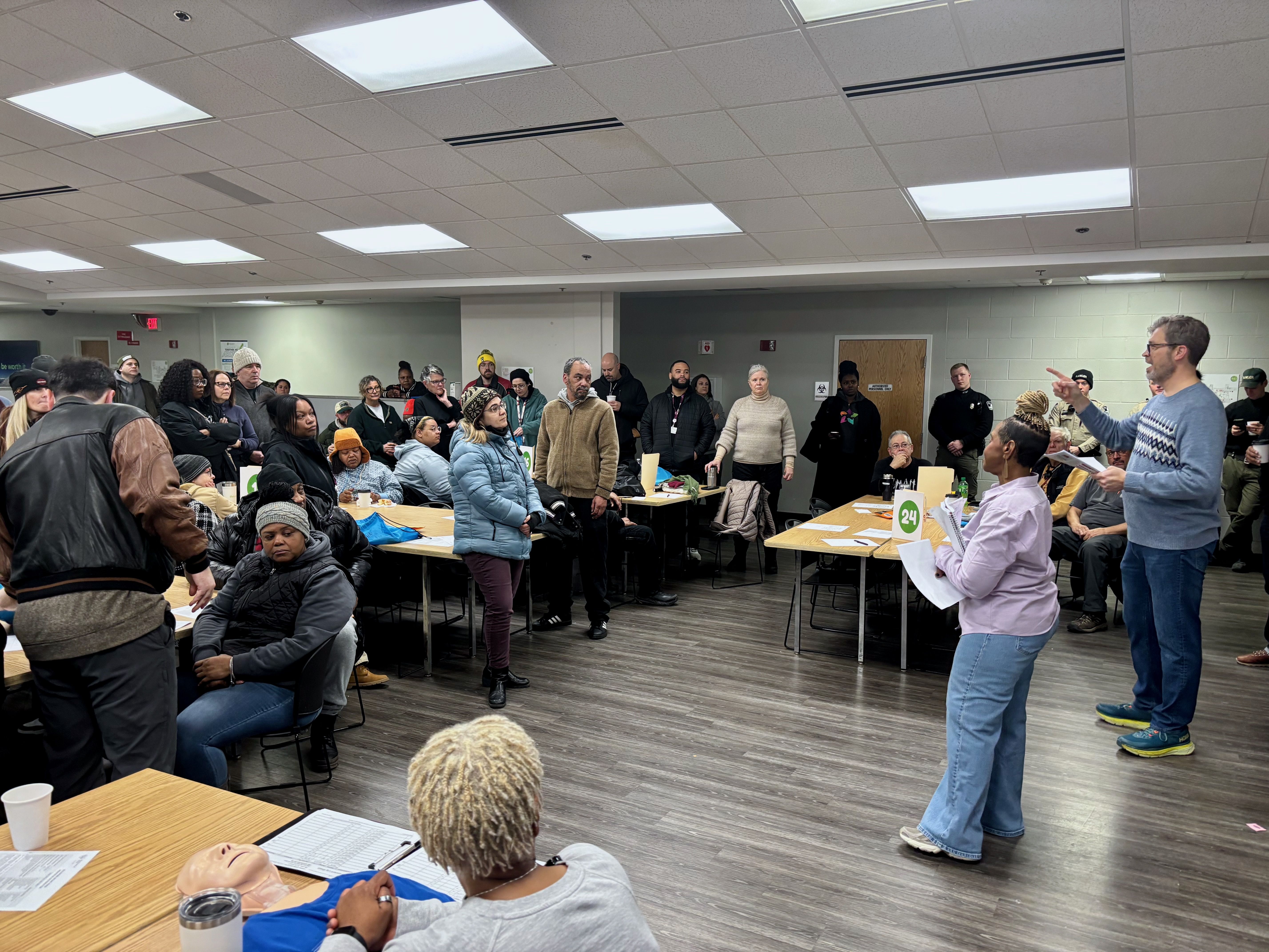 A group of people in winter clothing gather in a room, some seated at tables with papers, while a man and woman stand and speak, with others attentively listening around them.