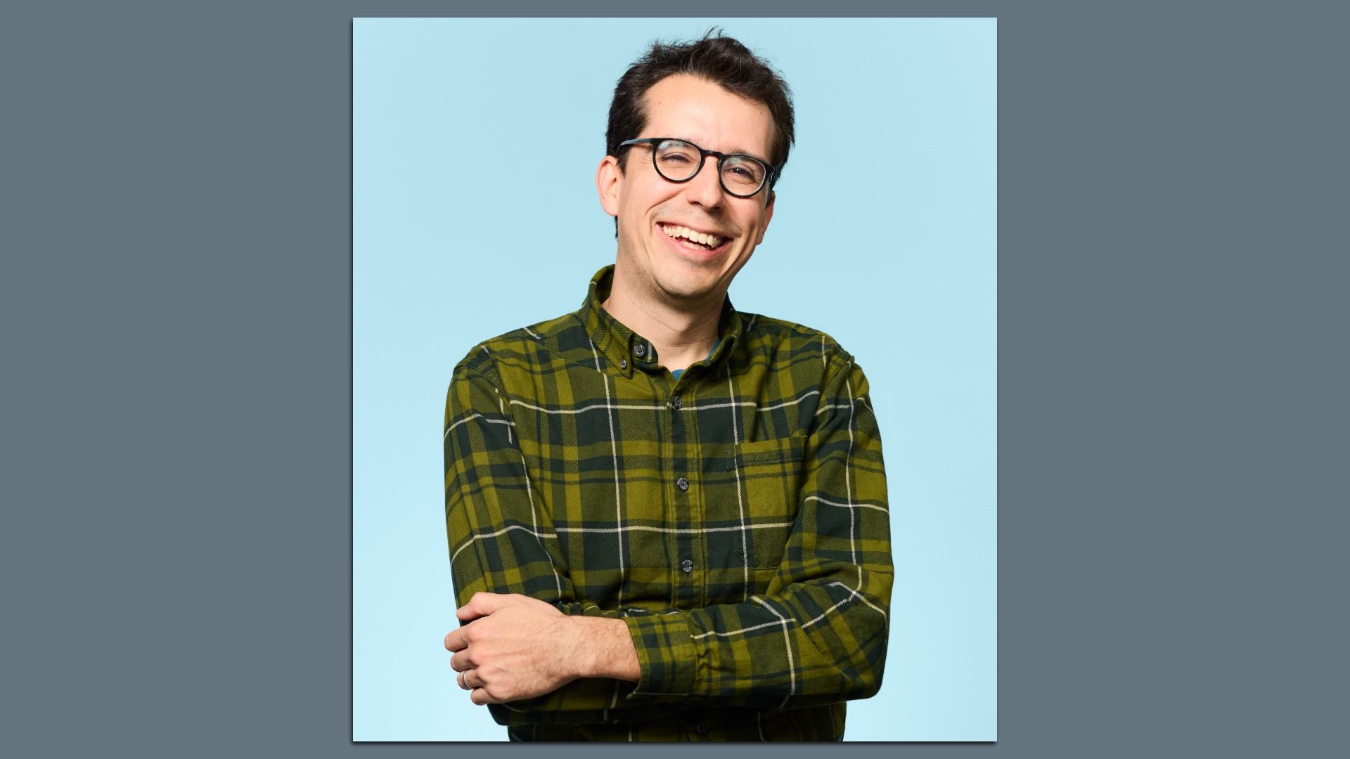 Portrait of Simon Hernandez-Arthur smiling with dark hair and round glasses, wearing a green plaid shirt, arms crossed, posing against a light blue studio background.