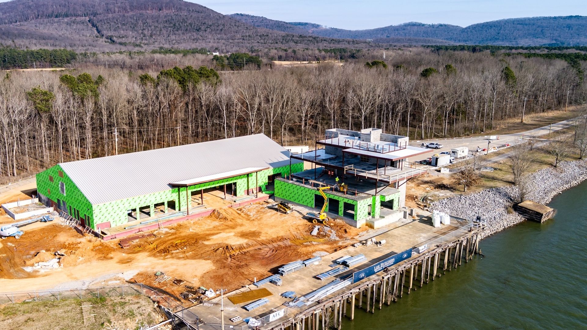 A riverfront construction site featuring a green-framed building with a white metal roof, dirt yard, heavy machinery, and a pier along the water; leafless trees and hills in the distance.