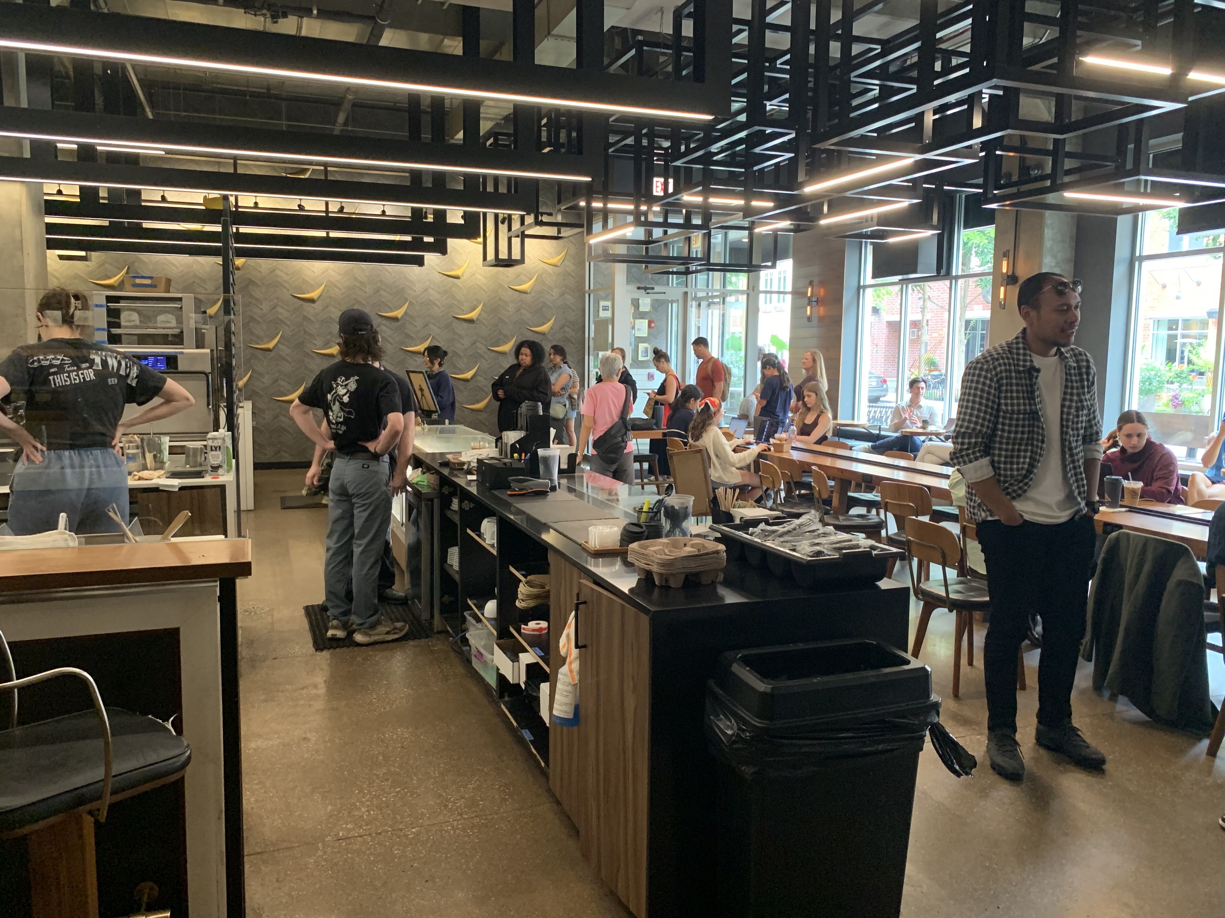 An industrial-style cafe with black metal ceiling beams and long lights. People queue at the counter while others sit at tables by large windows; a man in a plaid shirt stands at right.