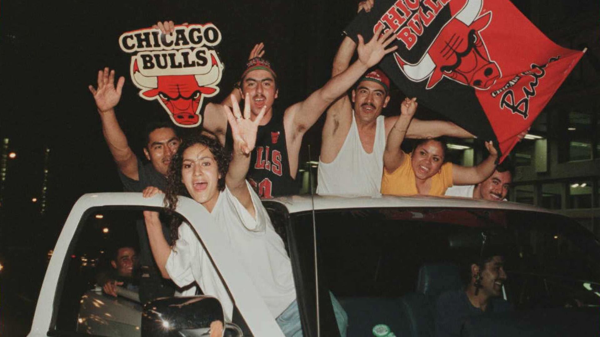 A group of excited fans ride atop a white pickup at night, waving. They hold Chicago Bulls banners with the red bull logo, wearing jerseys and bandanas.