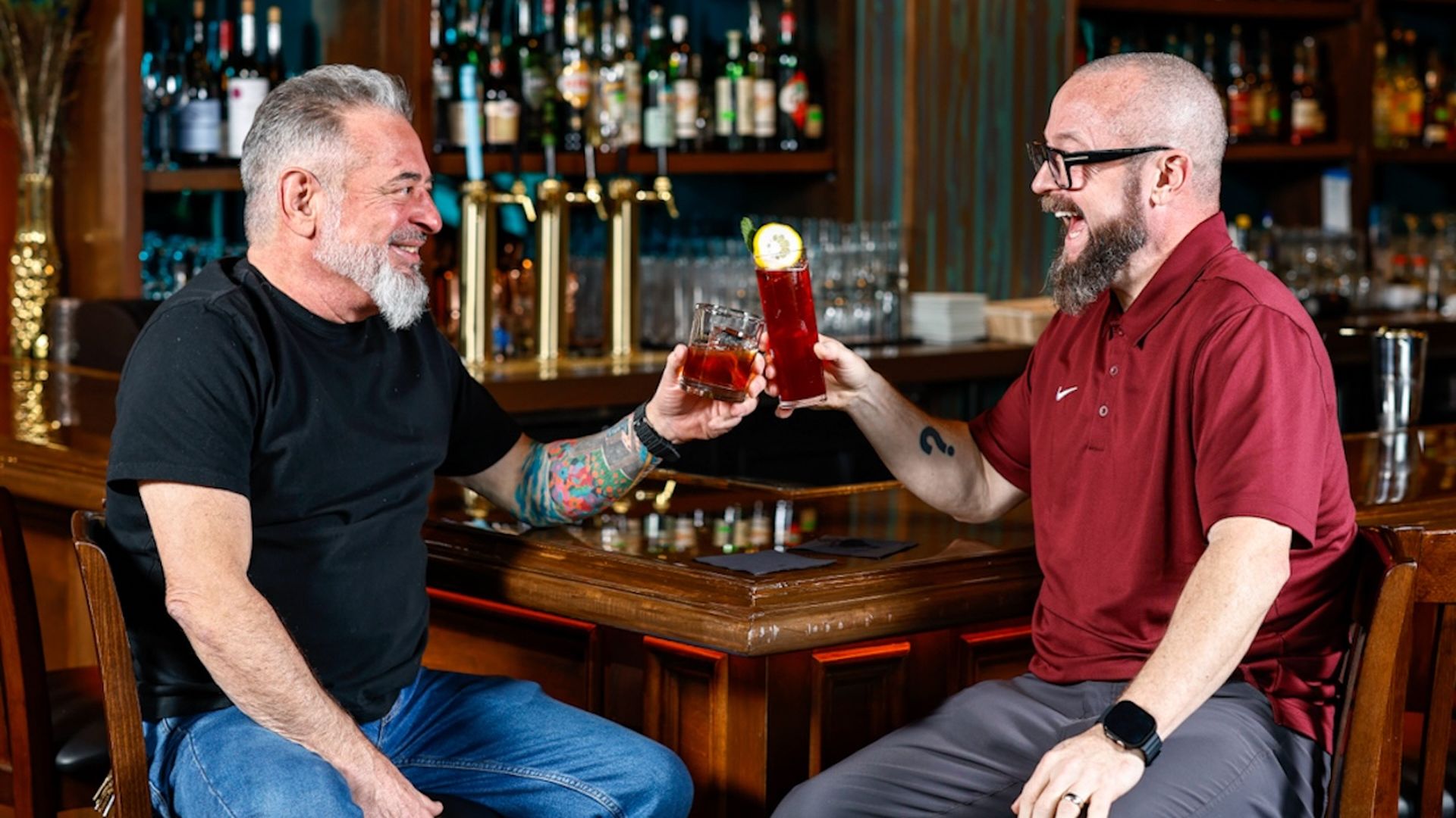 Two men sitting on stools at a wooden bar with shelves of liquor behind them, smiling and clinking their drinks, one in a black shirt with tattoos, the other in a red polo and colorful sneakers.
