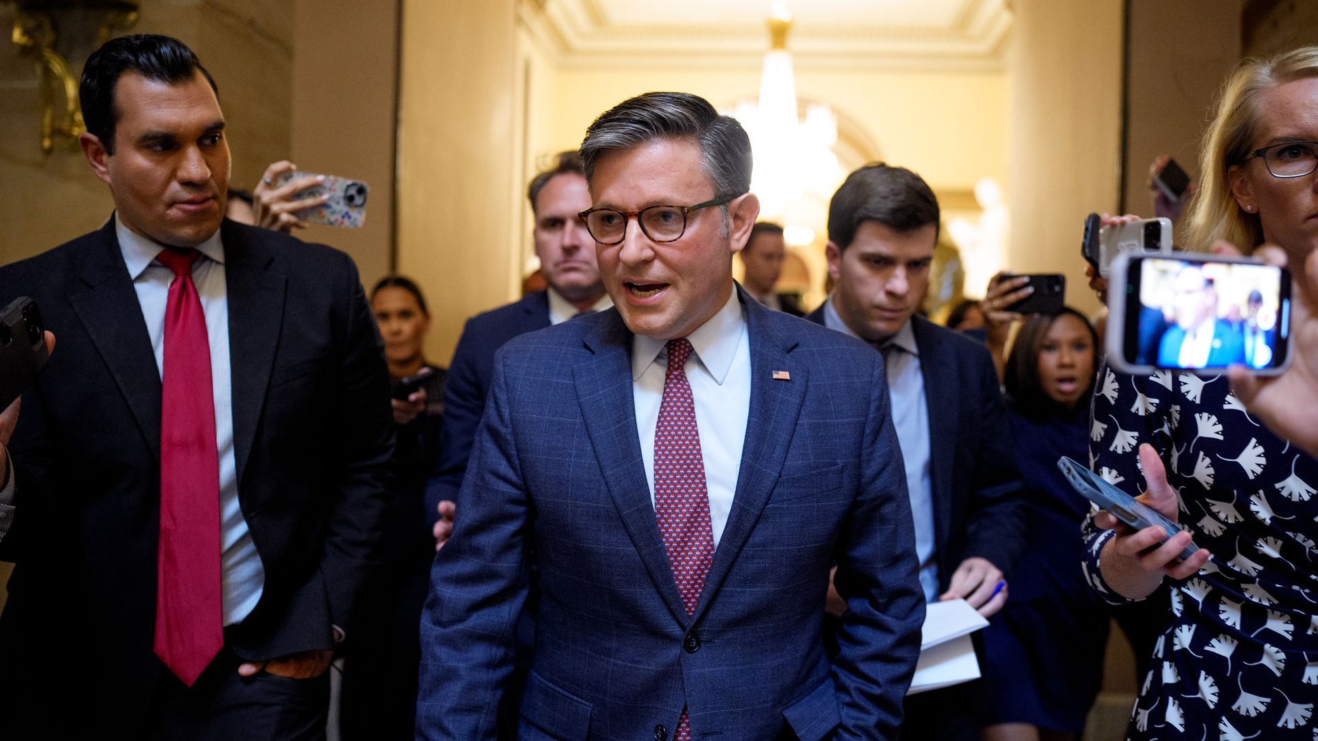 Man in a blue checkered suit and red tie speaking while walking, surrounded by people holding phones and recording him in a grand, well-lit hallway.