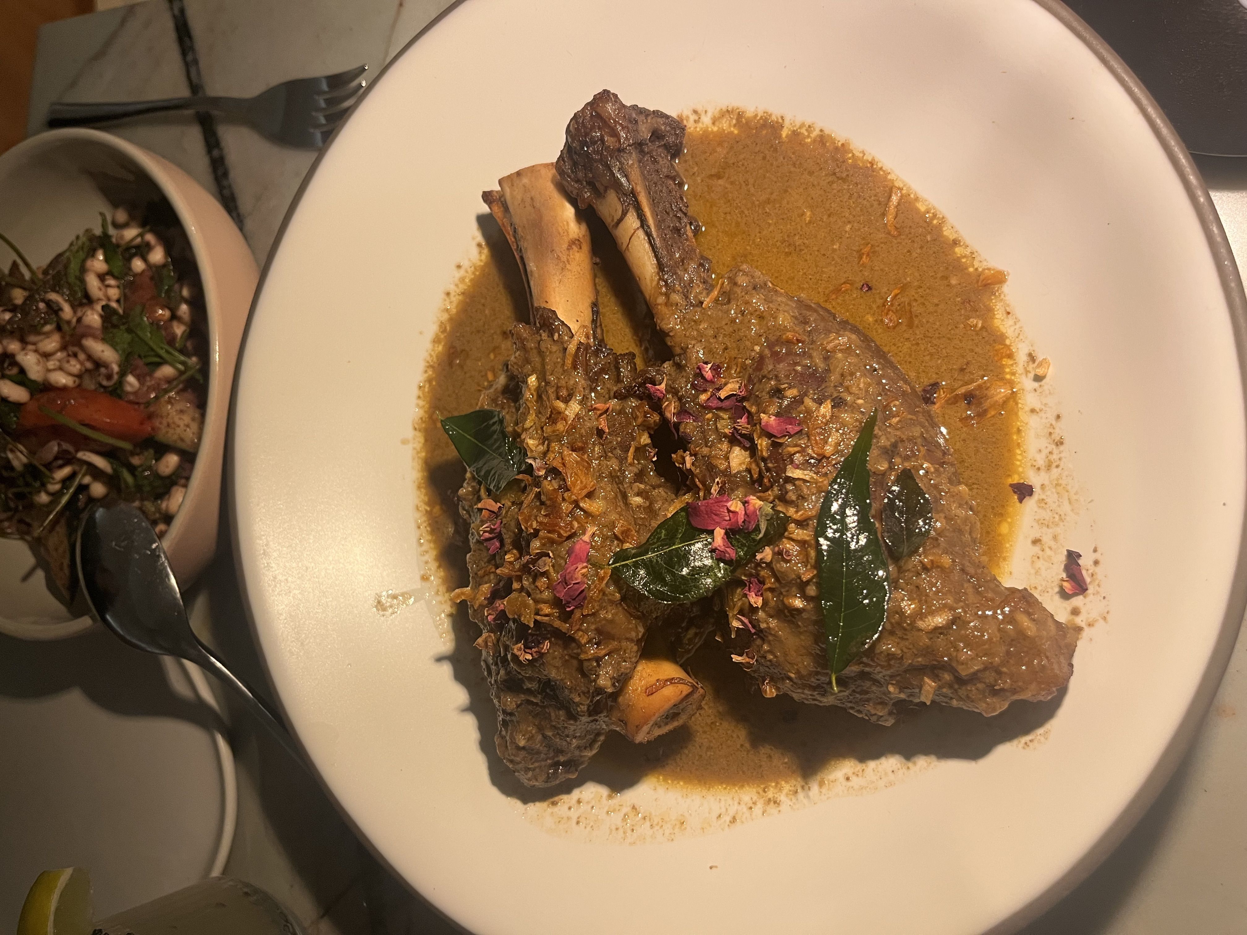 Braised bone-in meat in a yellow curry sauce on a white plate, topped with fried onions and pink petals; a side salad with greens and beans sits in a bowl nearby.