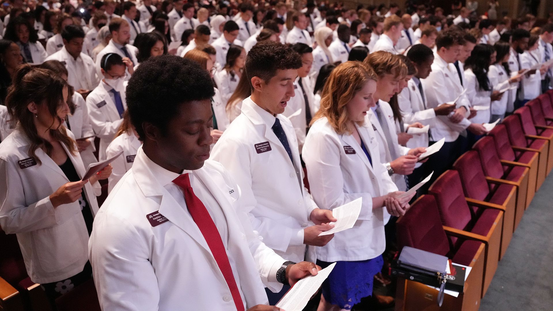 A group of medical students in white coats and ties stand in an auditorium, holding papers and reading aloud during a ceremony or event.
