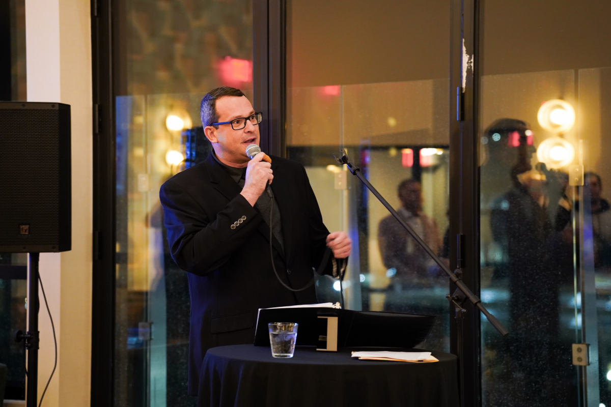 Matt Mandrella speaks into a microphone in front of a darkened window. 