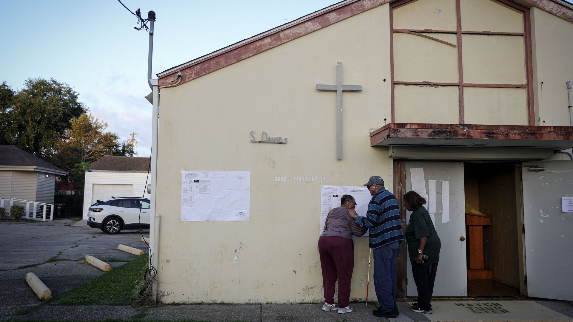 Three people huddle around a sample ballot poster outside of a church doorway.