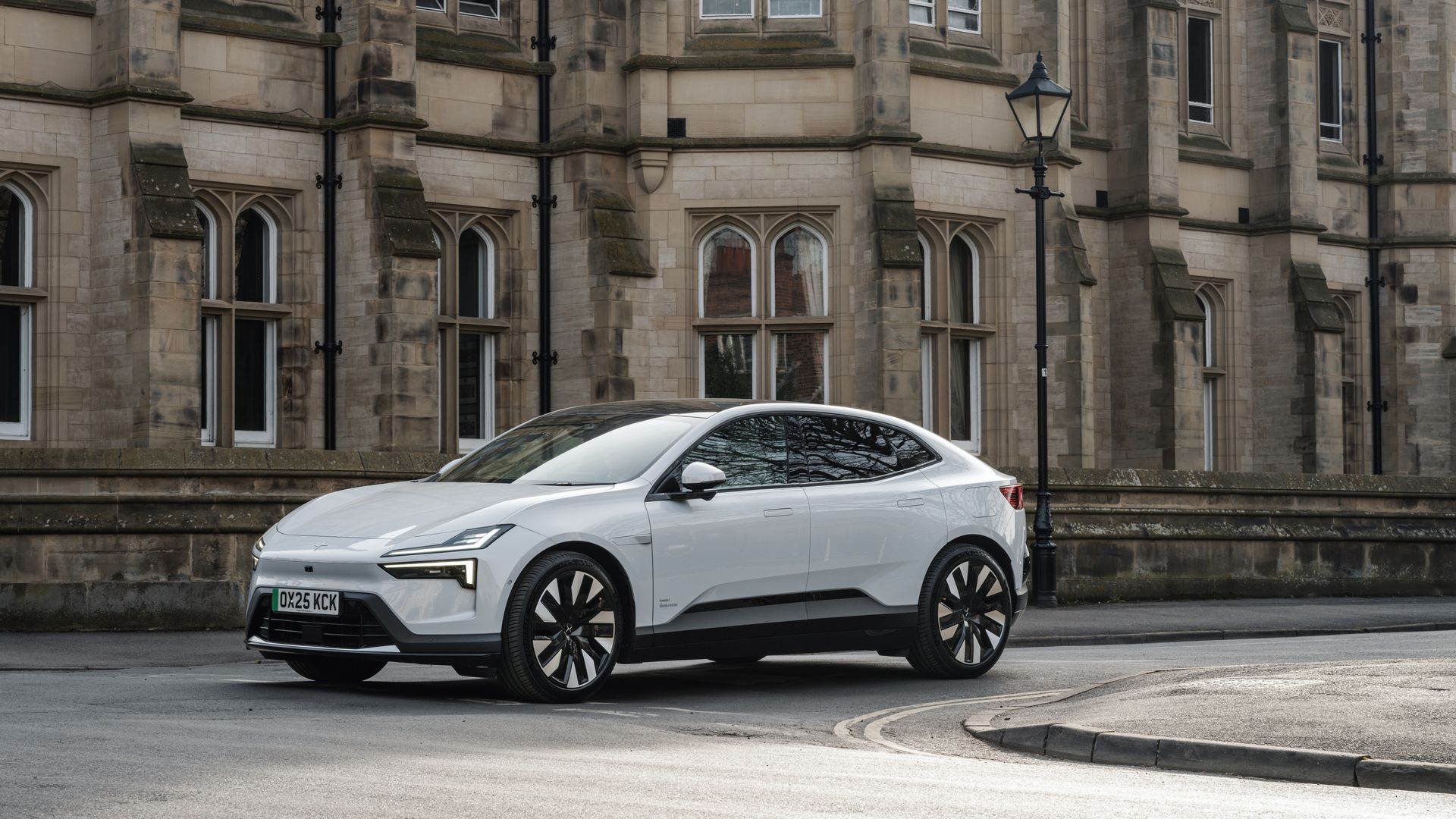 A sleek white electric Polestar 4 parked along a city street in front of a historic beige stone building with arched windows and a classic lamppost.