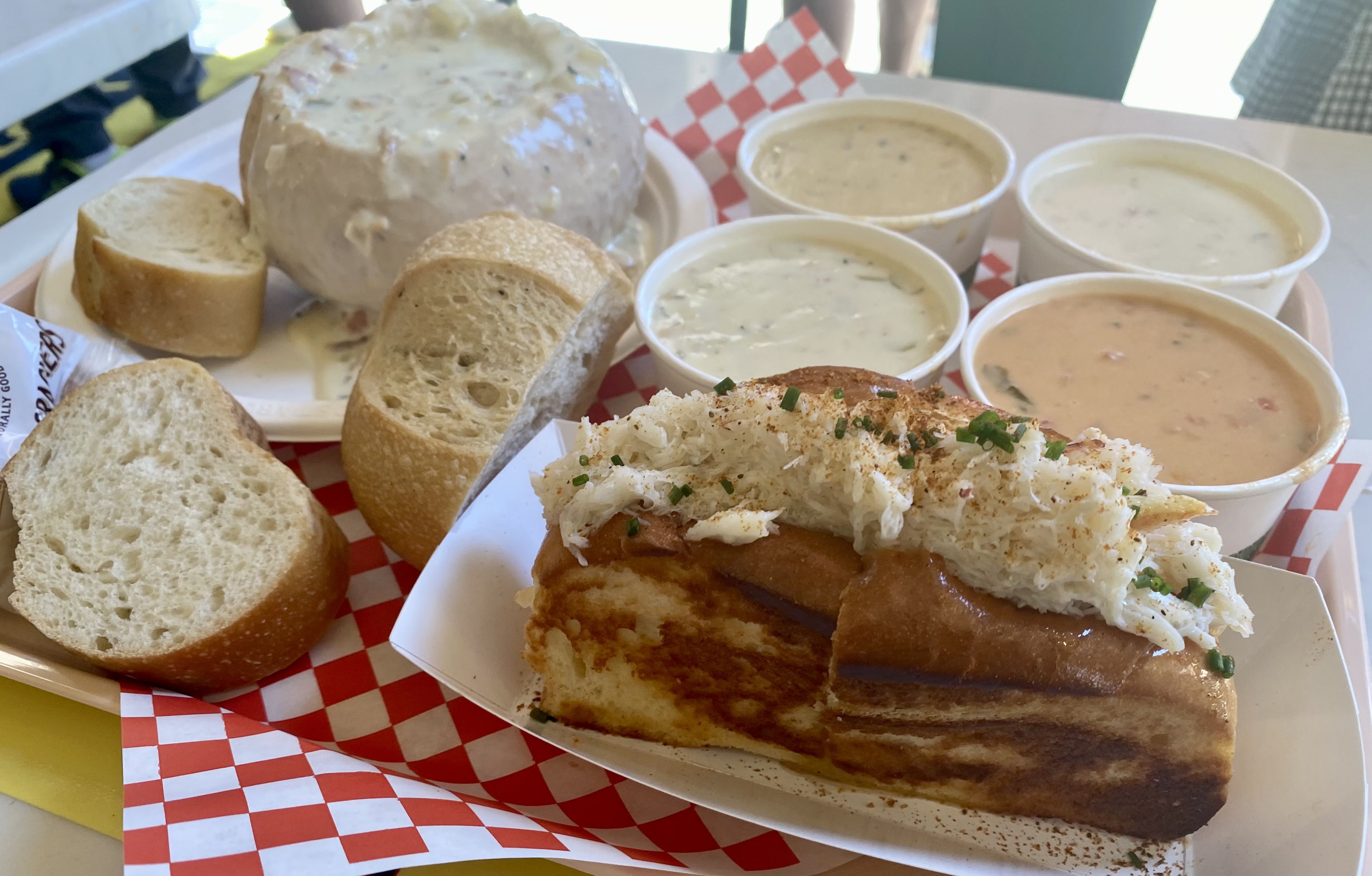 Four paper bowls of chowder next to a crab roll and a bread bowl filled with chowder, on a tray. 