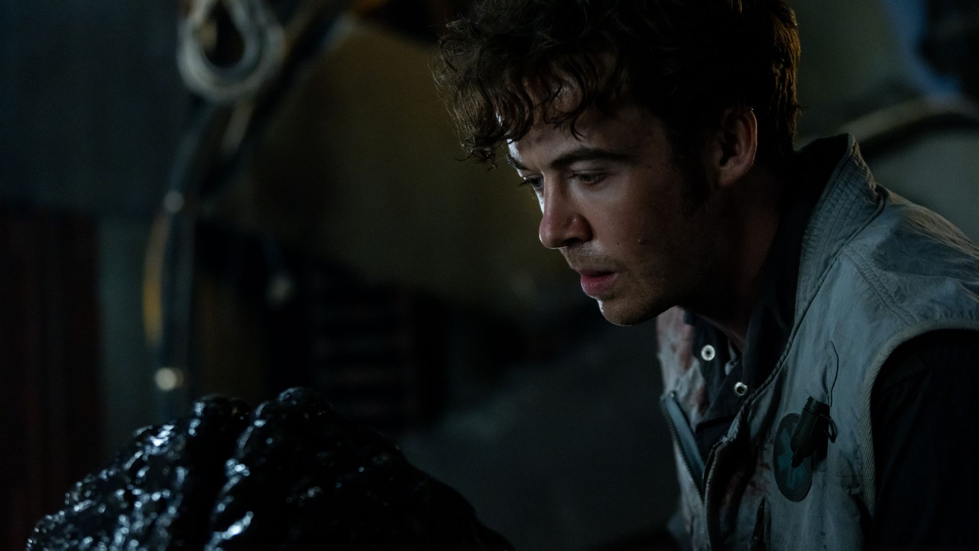 A young man with curly hair wearing a gray vest looks intently at a shiny black object in a dimly lit room with industrial background elements.