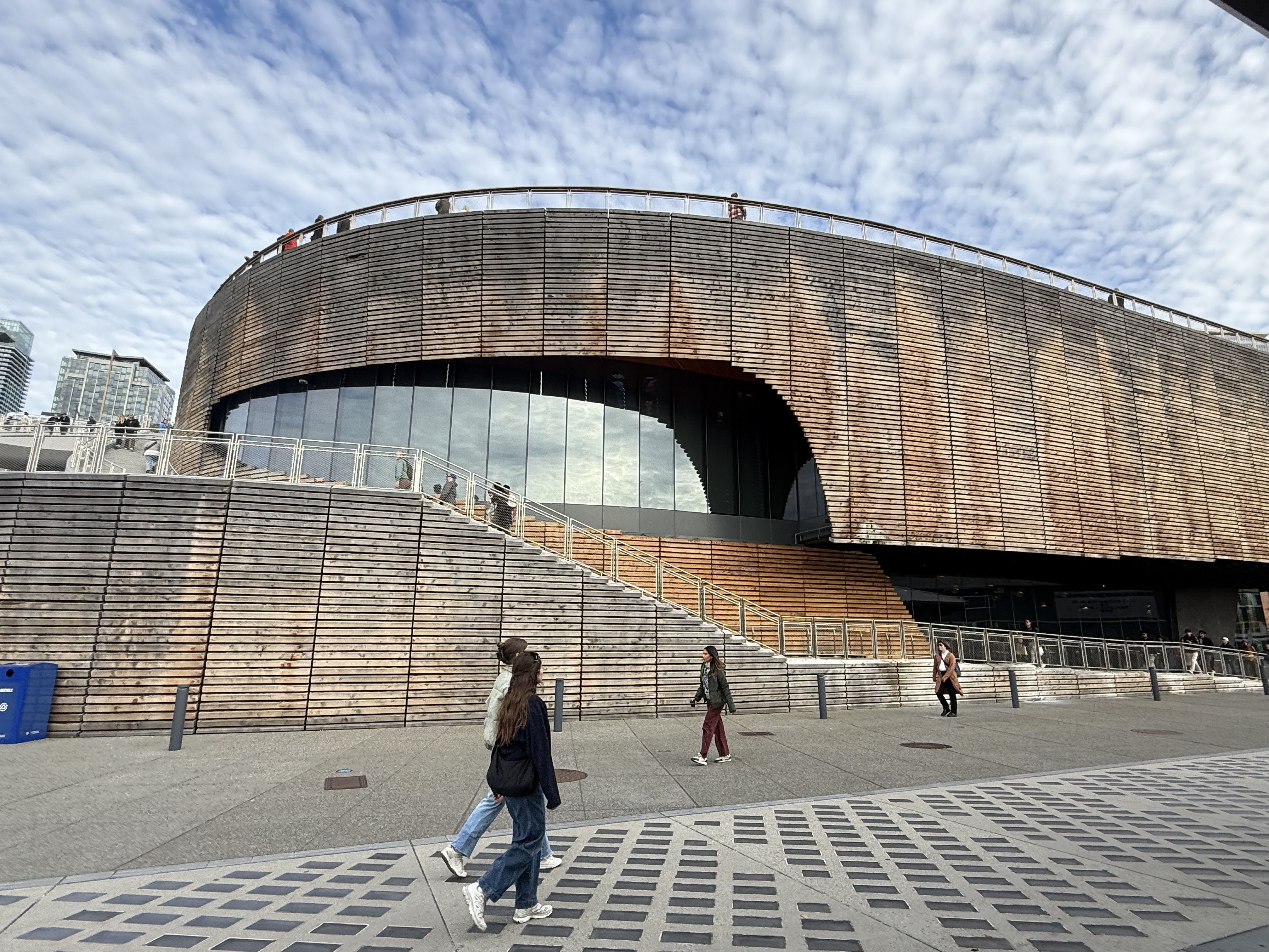 Curved modern building with wooden slats and large glass window, people walking outside on gray pavement with geometric designs, and a partly cloudy sky overhead.