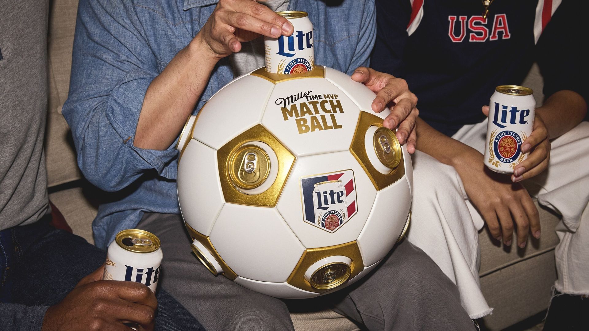 Two hands and several Miller Lite cans surround a white-and-gold MVP soccer ball labeled MATCH BALL; one hand places a can atop the ball, while a person in a USA shirt holds another can nearby.