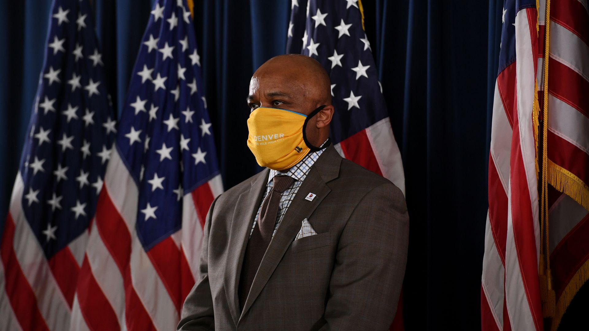 A photo of Denver Mayor Michael Hancock wearing a mask and standing in front of several American flags.