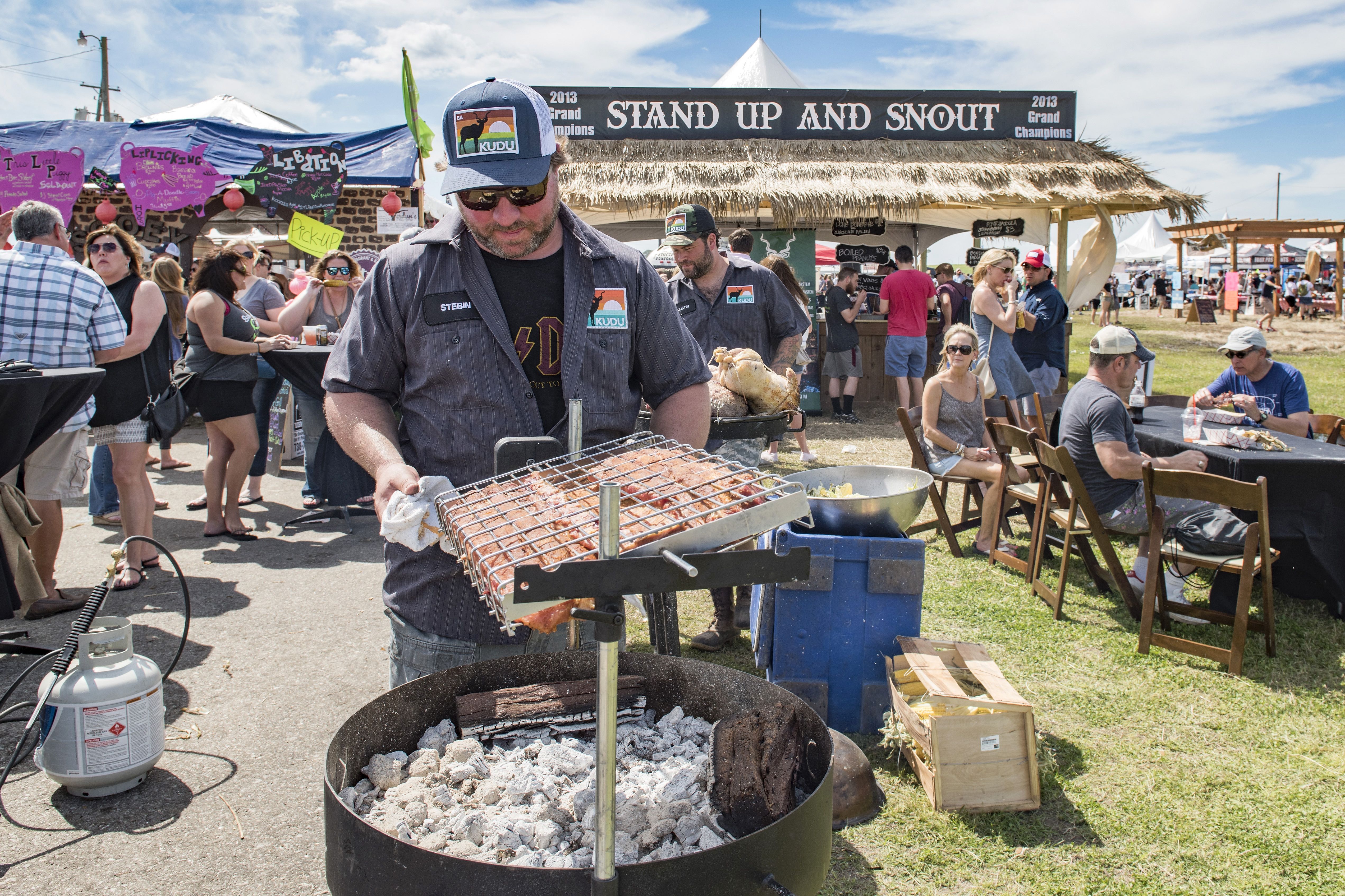 A man adjusts some meat over hot coals in the middle of a festival. Behind him is a tent that says "Stand up and snout."