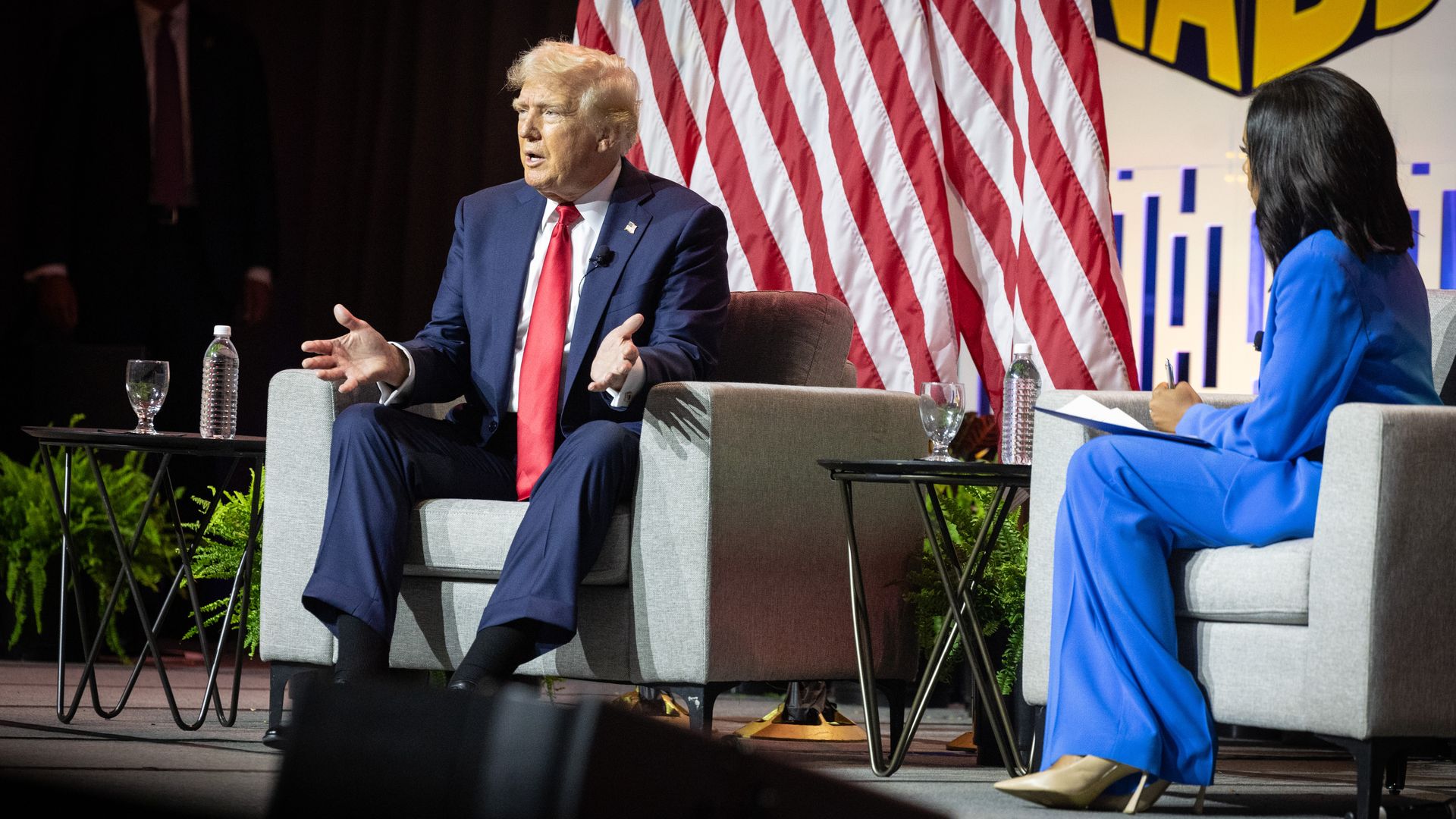 Donald Trump, wearing a blue suit, sits in a white chair next to Rachel Scott, wearing a light blue suit, also sitting in a white chair, surrounded by tables with water on them, a white backdrop and an American flag.