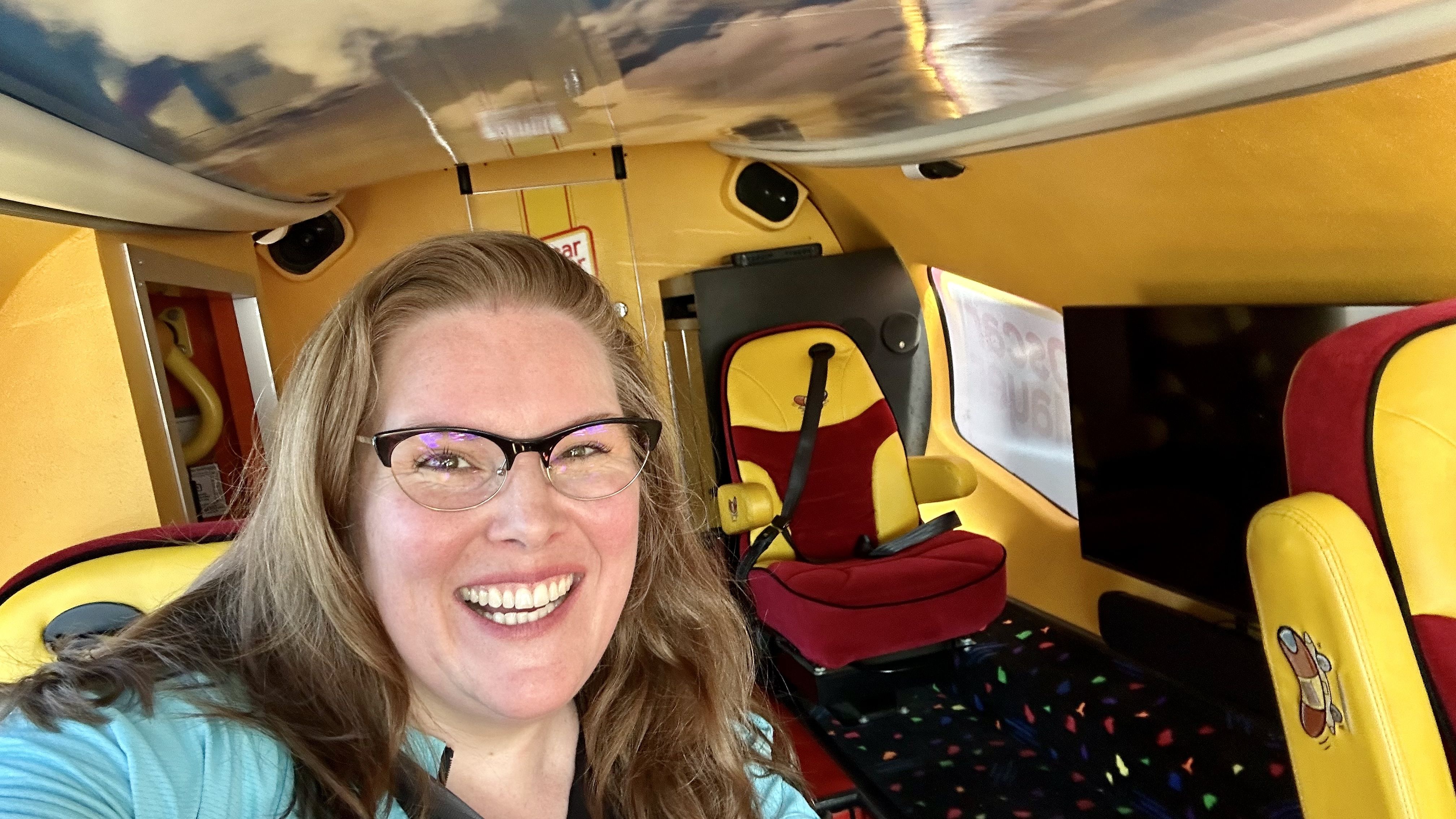 Smiling woman with glasses inside the Wienermobile, wearing a teal top. Red seats with cartoon designs; cabin interior and painted ceiling visible above.