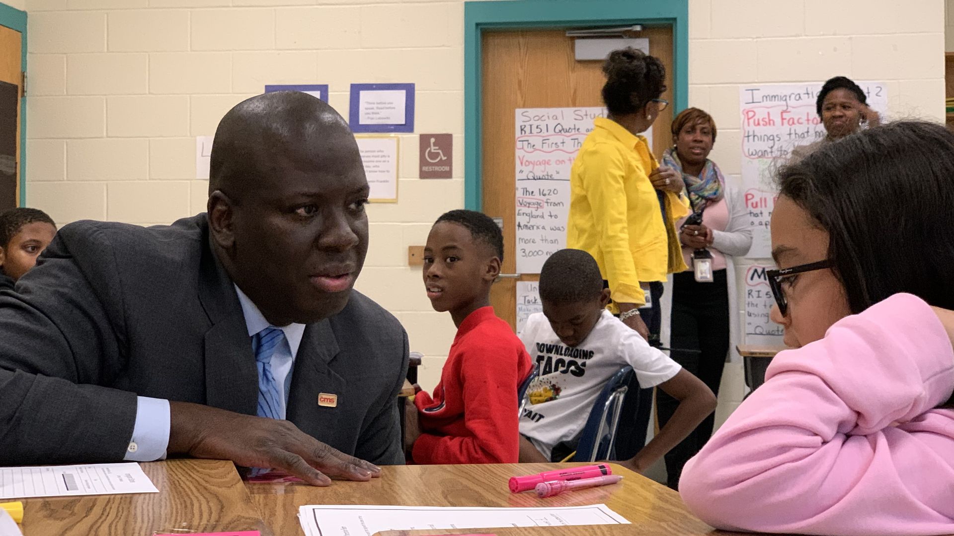 Superintendent Winston sits inside a classroom at a desk, talking to child. 