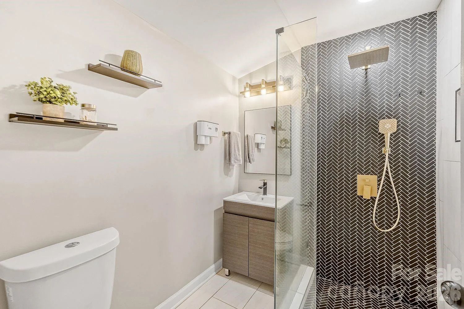 Modern bathroom with white walls, a white toilet, and a wooden vanity with a sink and mirror. The shower has black herringbone tiles and gold fixtures, including a rainfall showerhead.