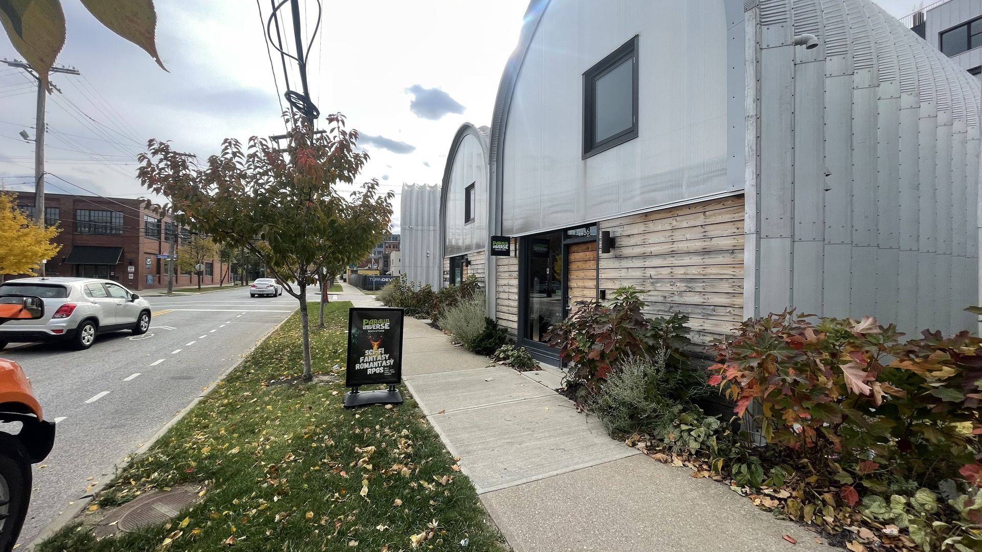 Sidewalk view of a street with silver arched buildings with wooden accents, small trees with fall-colored leaves, a black sign reading Parallel Universe bookstore with sci-fi, fantasy, romantic, RPGs text.
