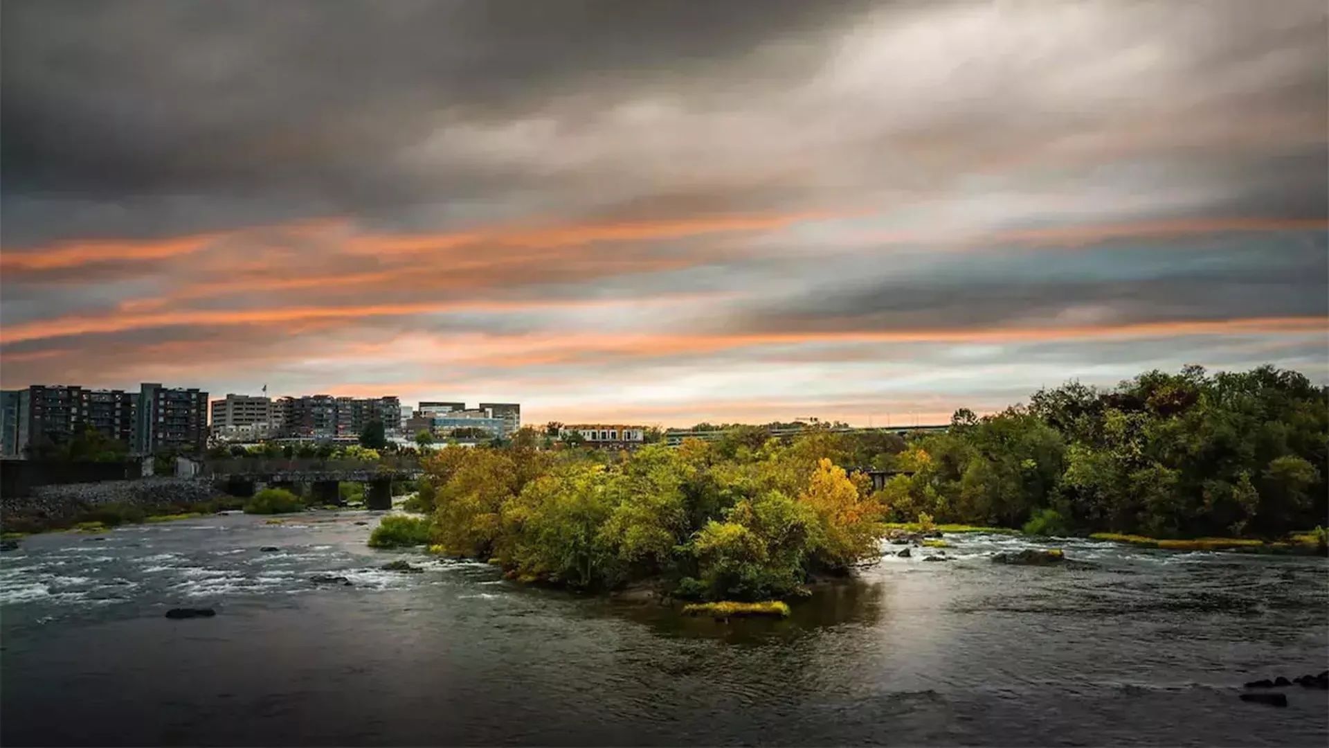 Sunset over a river with a city on the left and trees on the right; orange-pink clouds glow across a moody sky, with gentle ripple water in the foreground.