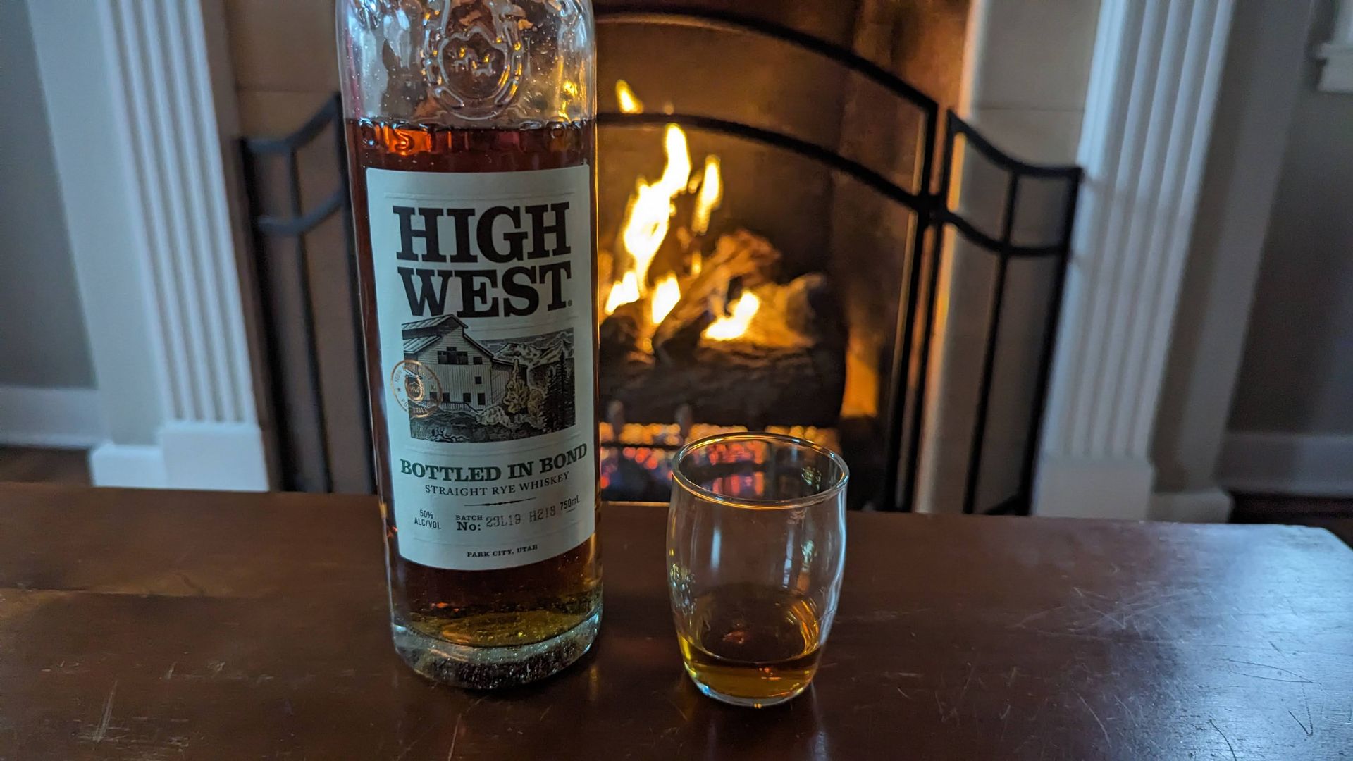 A bottle of High West Bottled in Bond rye whiskey next to a glass of whiskey in front of a fireplace.