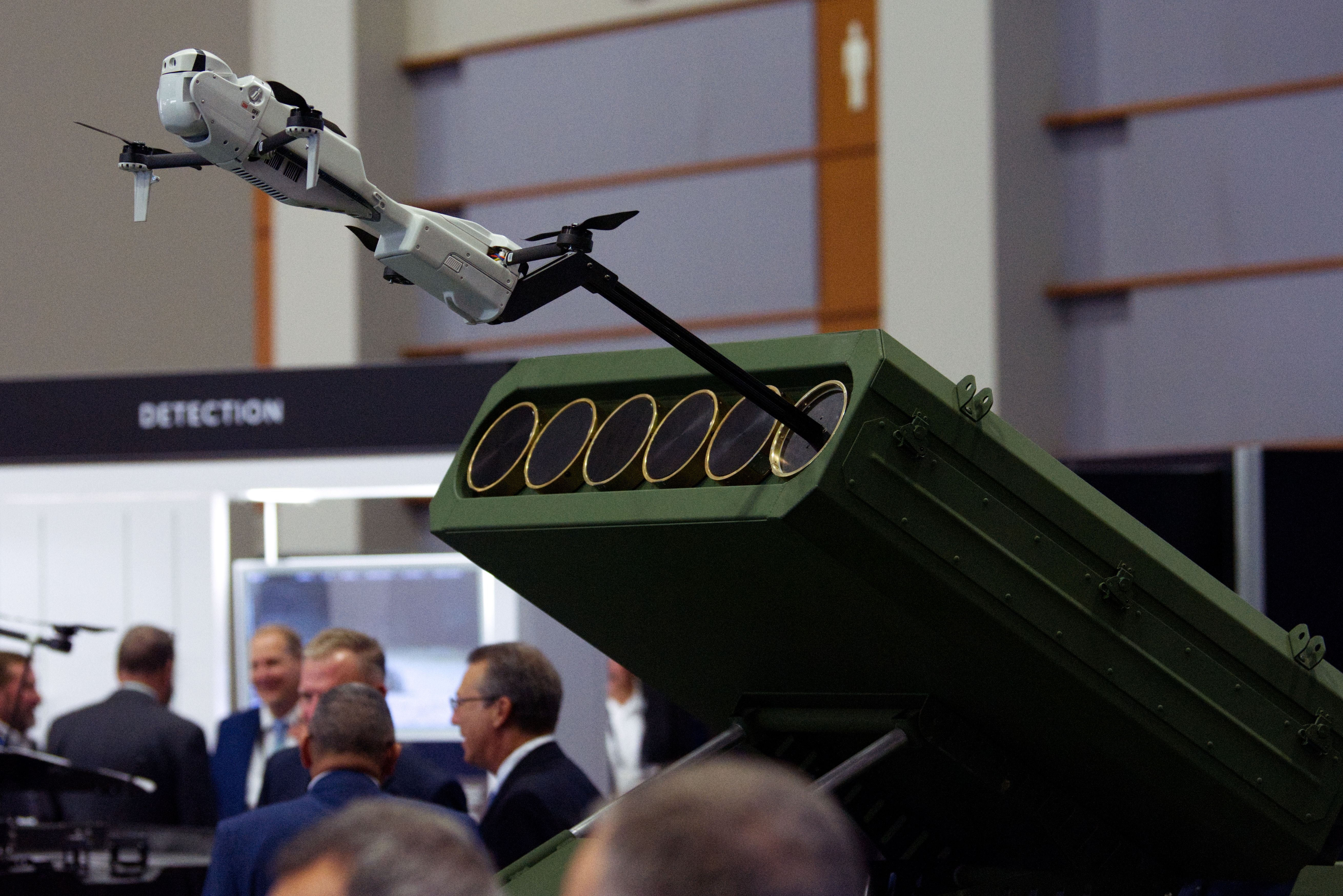 A Rogue 1 drone pops out of a launcher at the Teledyne booth at AUSA. The drone is grey. The launcher is green. People can be seen mingling in the background.