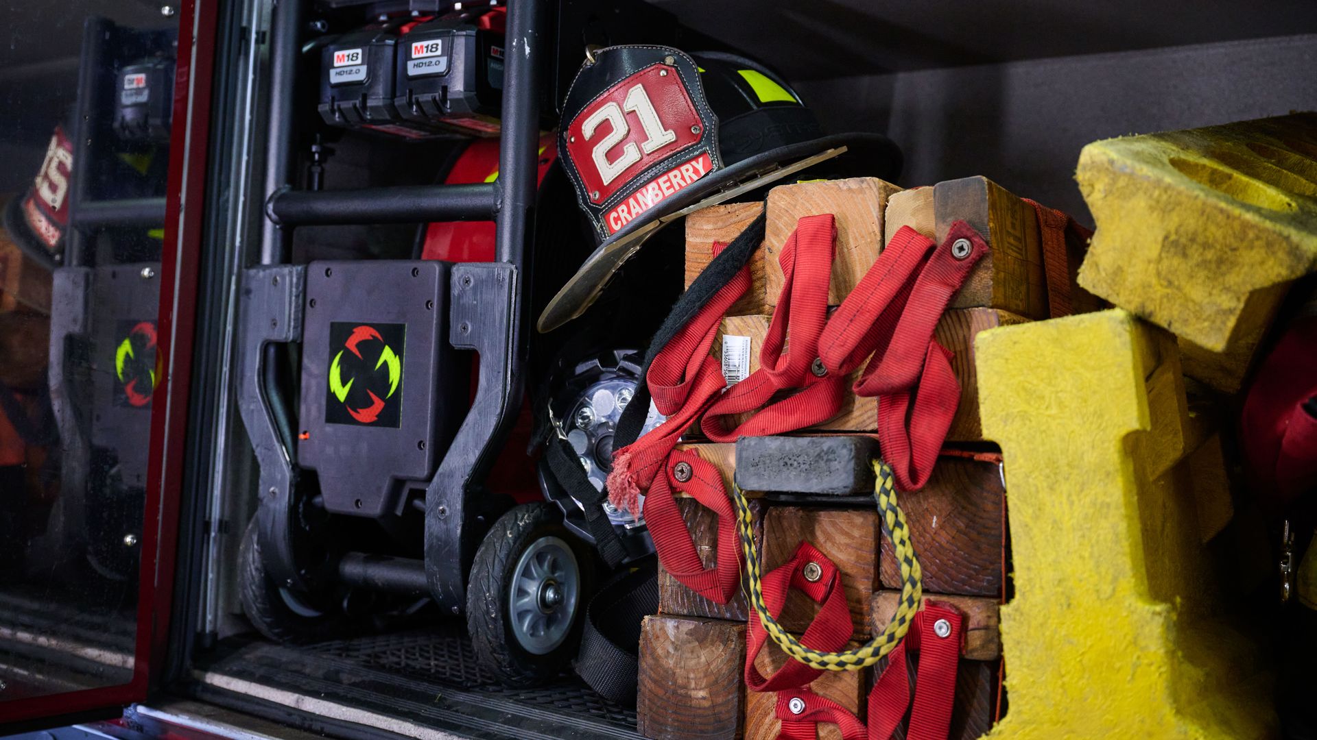 Inside a fire truck storage bay, a black firefighter helmet labeled 21 with the name CRANBERRY rests on wooden blocks amid red straps, yellow foam, and M18 battery packs.