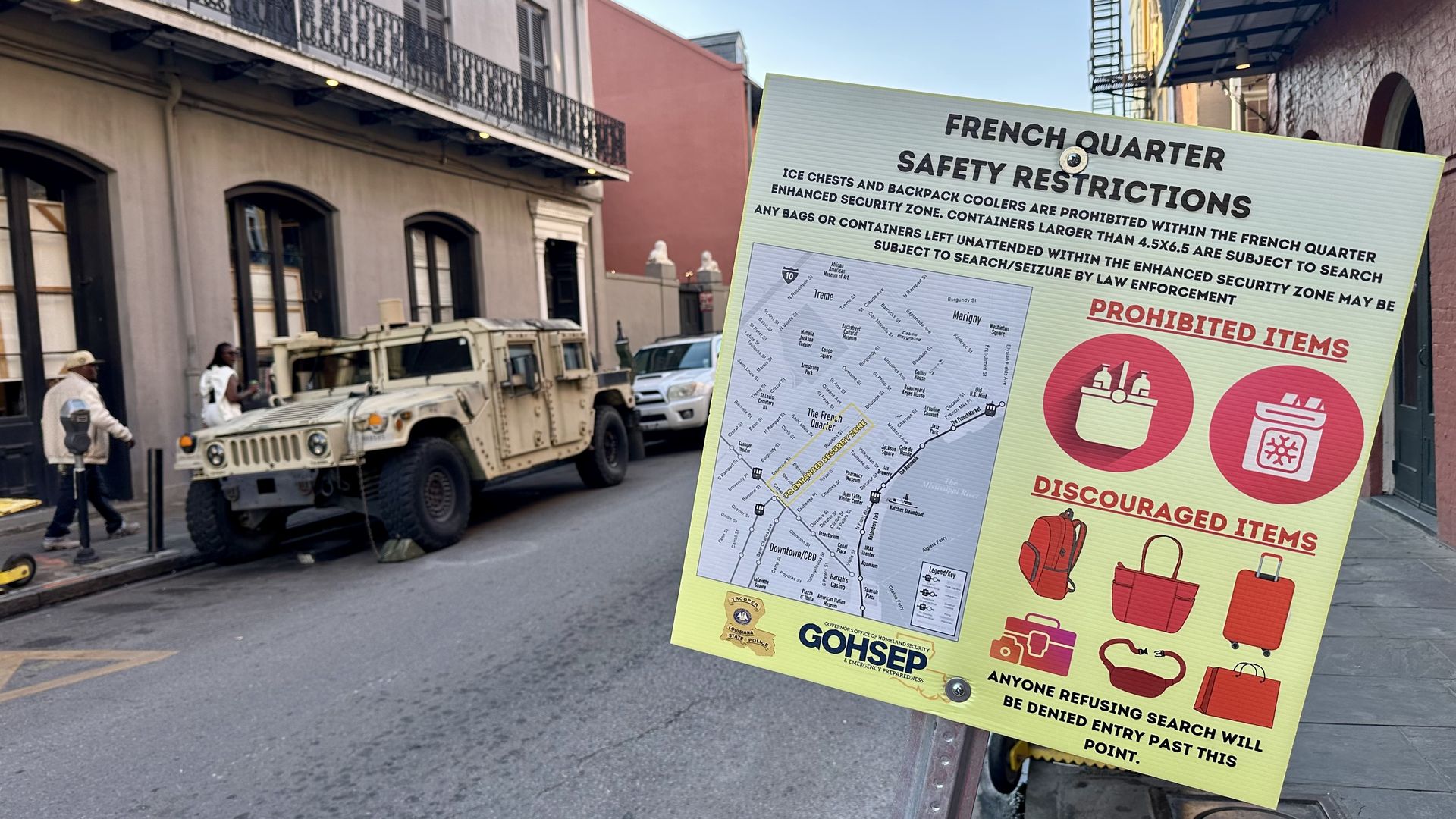 Yellow French Quarter safety restrictions sign listing banned coolers and backpacks, with prohibited and discouraged items icons, set on a street with parked vehicles and pedestrians.