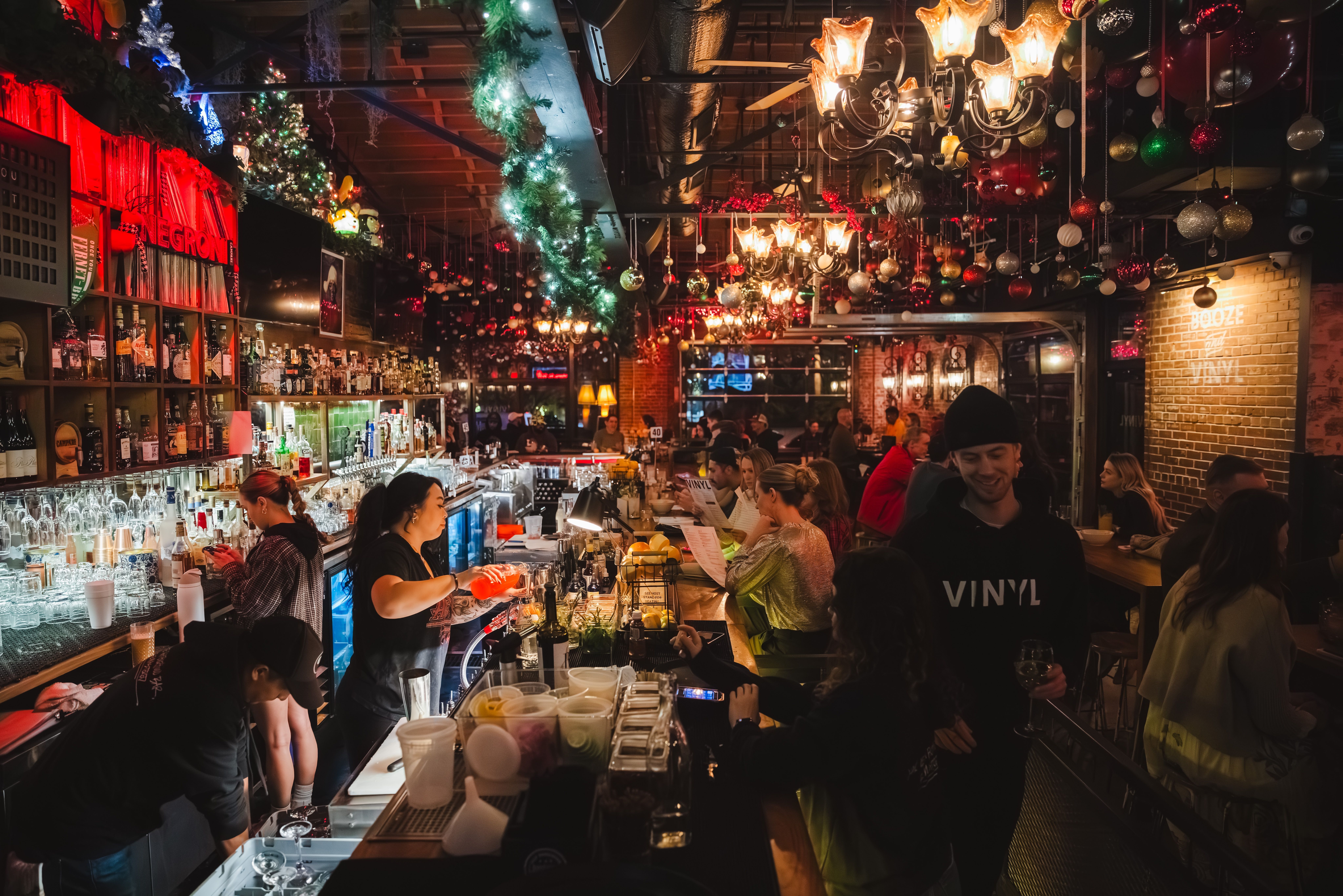 Cozy bar with festive Christmas decorations, green garlands, and red, gold ornaments. Patrons sit and chat while bartenders prepare drinks under warm chandelier lighting.