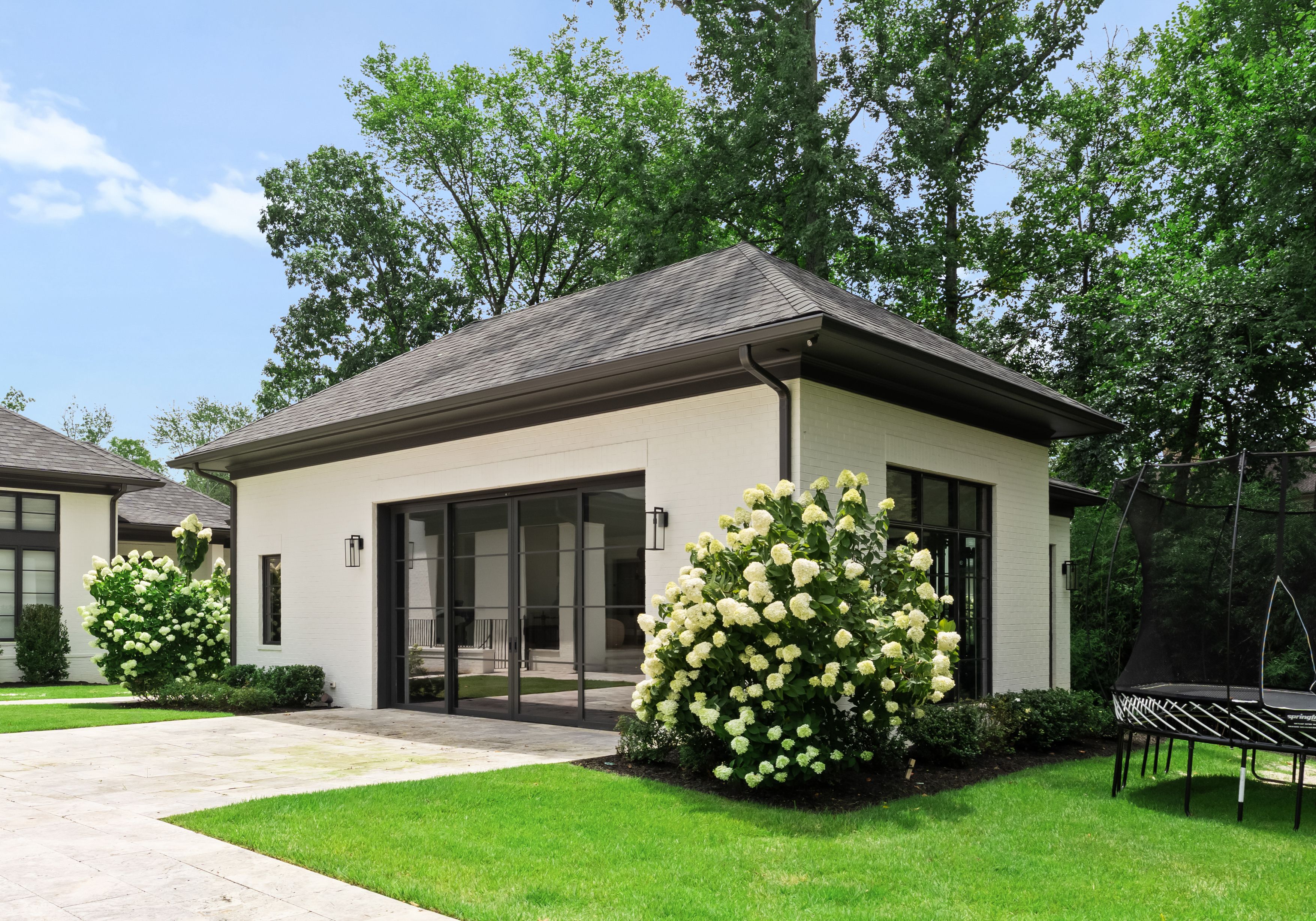 White brick house with dark roof and large black-framed glass doors, surrounded by green lawns, blooming white hydrangea bushes, and tall trees under a partly cloudy blue sky.
