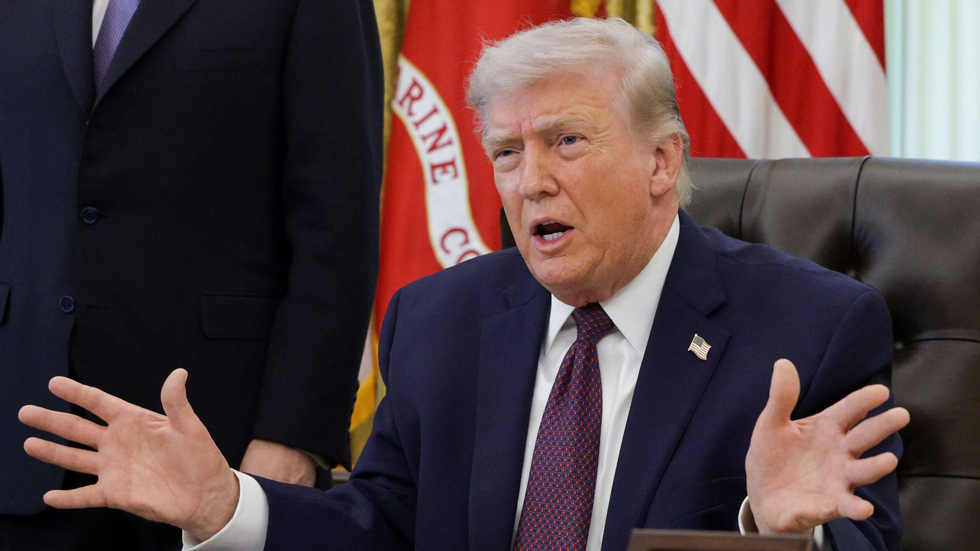 President Trump, an older man in a dark navy suit and red patterned tie sits at a desk, hands spread as he speaks. Behind him are American flags and a gold-framed emblem in a formal office setting.