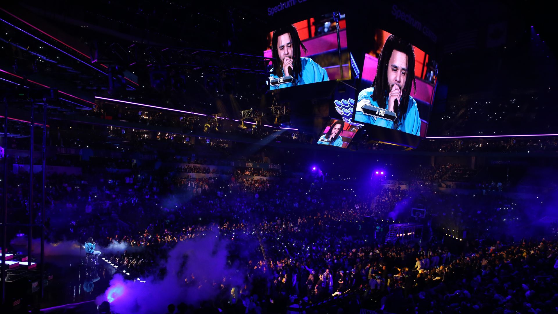 A large indoor arena concert with a singer on a circular central screen wearing a blue shirt and dreadlocks, microphone in hand; purple lights, smoke, and a packed crowd.