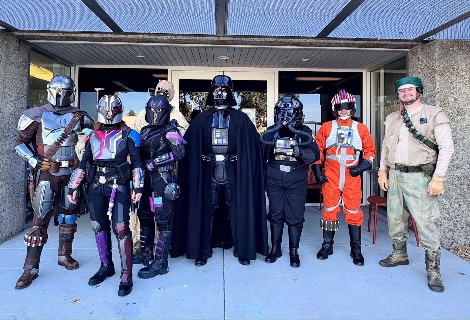 Group of cosplayers dressed as Mandalorians, with Darth Vader in the center, plus an orange-suited figure and a beige-clad man, posing outside a building under a glass canopy.