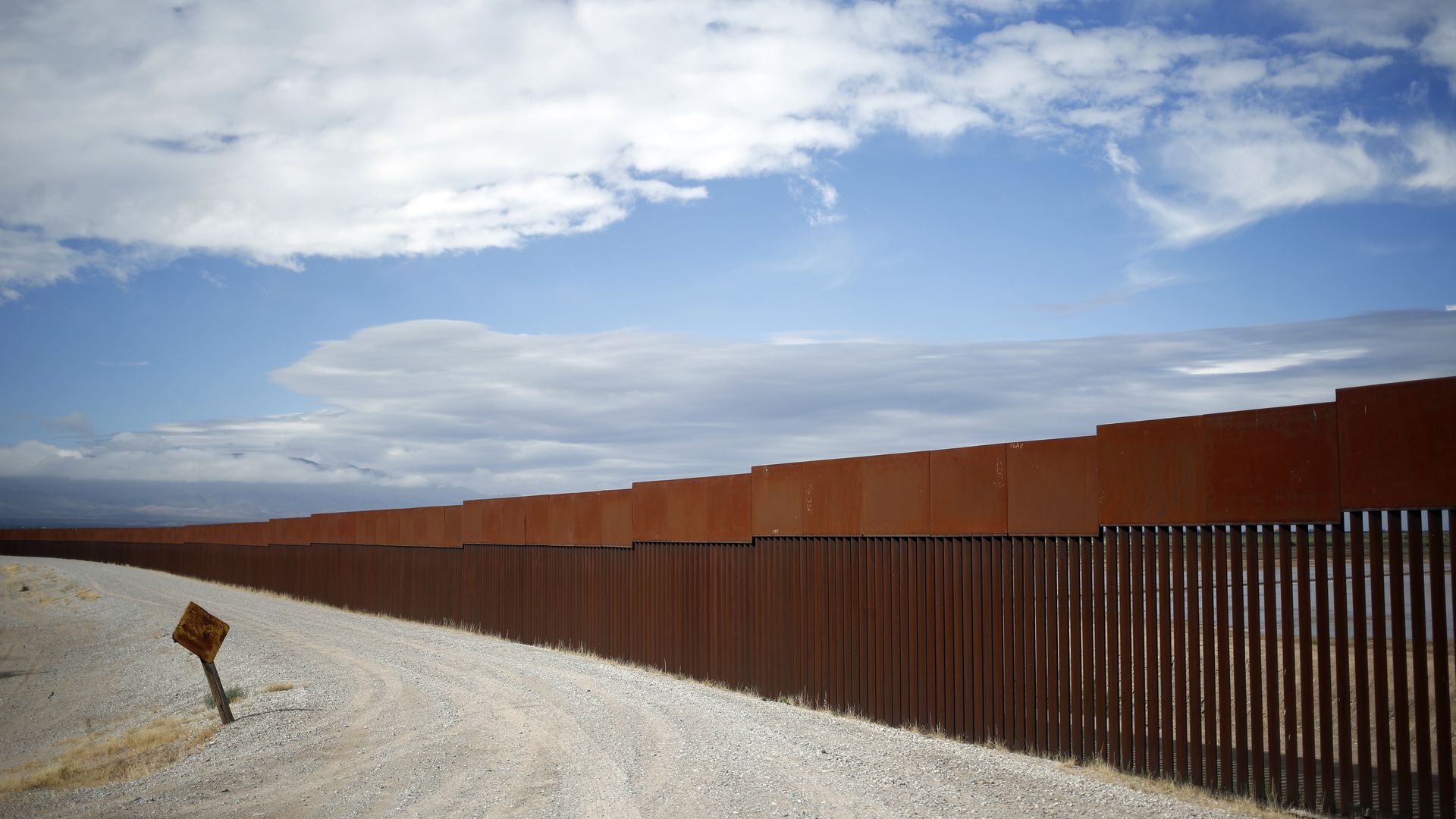 Long rust-colored metal border fence alongside a dirt road with dry grass under a partly cloudy blue sky.