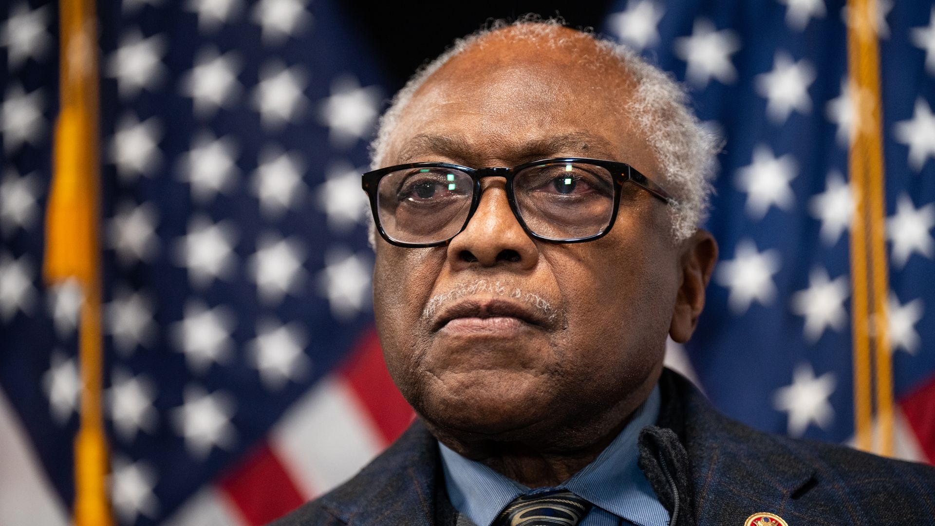 Rep. Jim Clyburn, wearing a dark blue suit, blue shirt and glasses, standing in front of American flags.