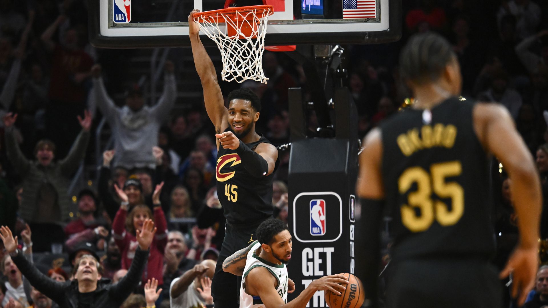 Donovan Mitchell, in black jersey, winks and points at camera while hanging from basketball hoop with one hand