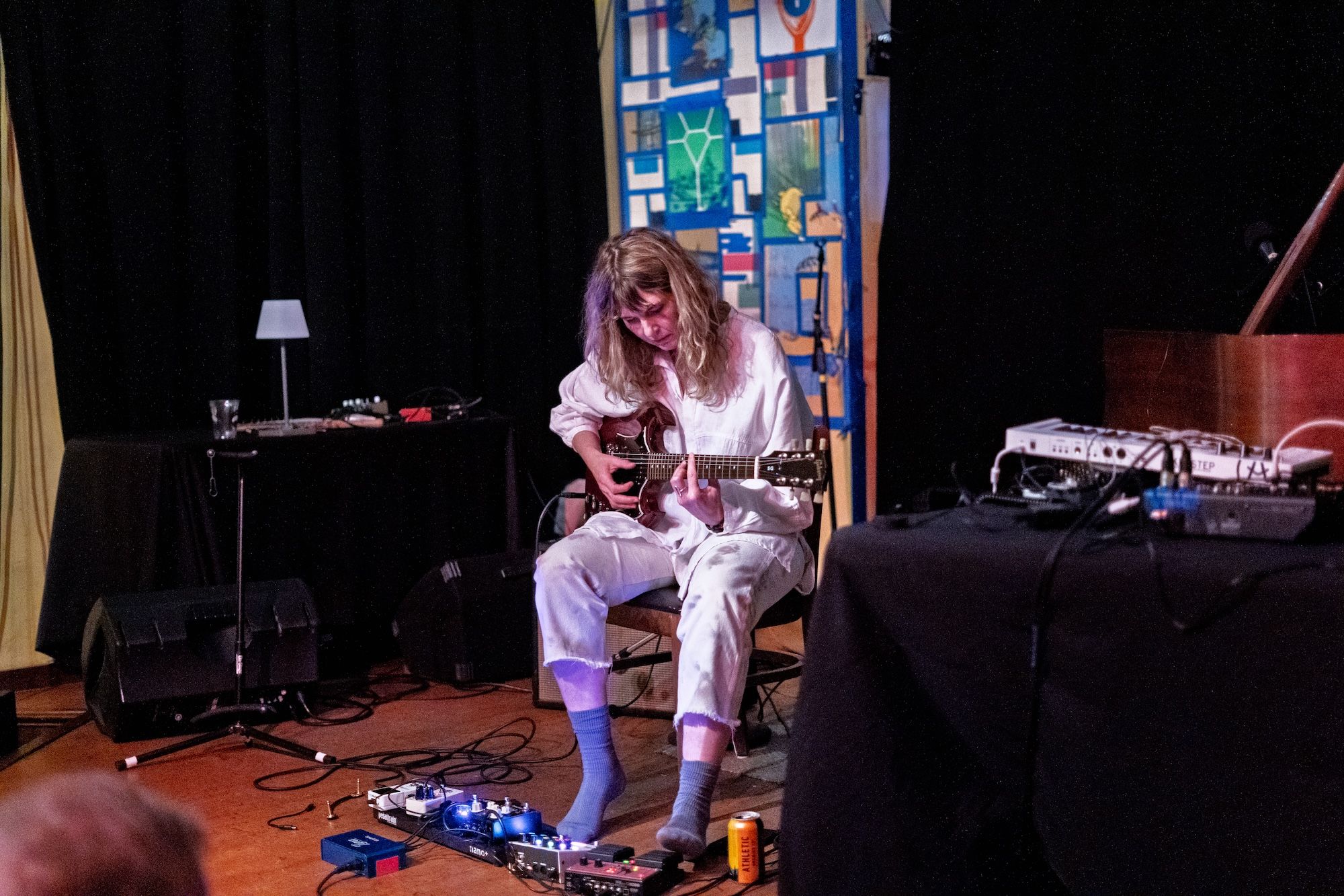 Female guitarist in white, seated center stage, playing an electric guitar. A blue collage backdrop rises behind her; pedals, cables, and stage gear spread across the floor.
