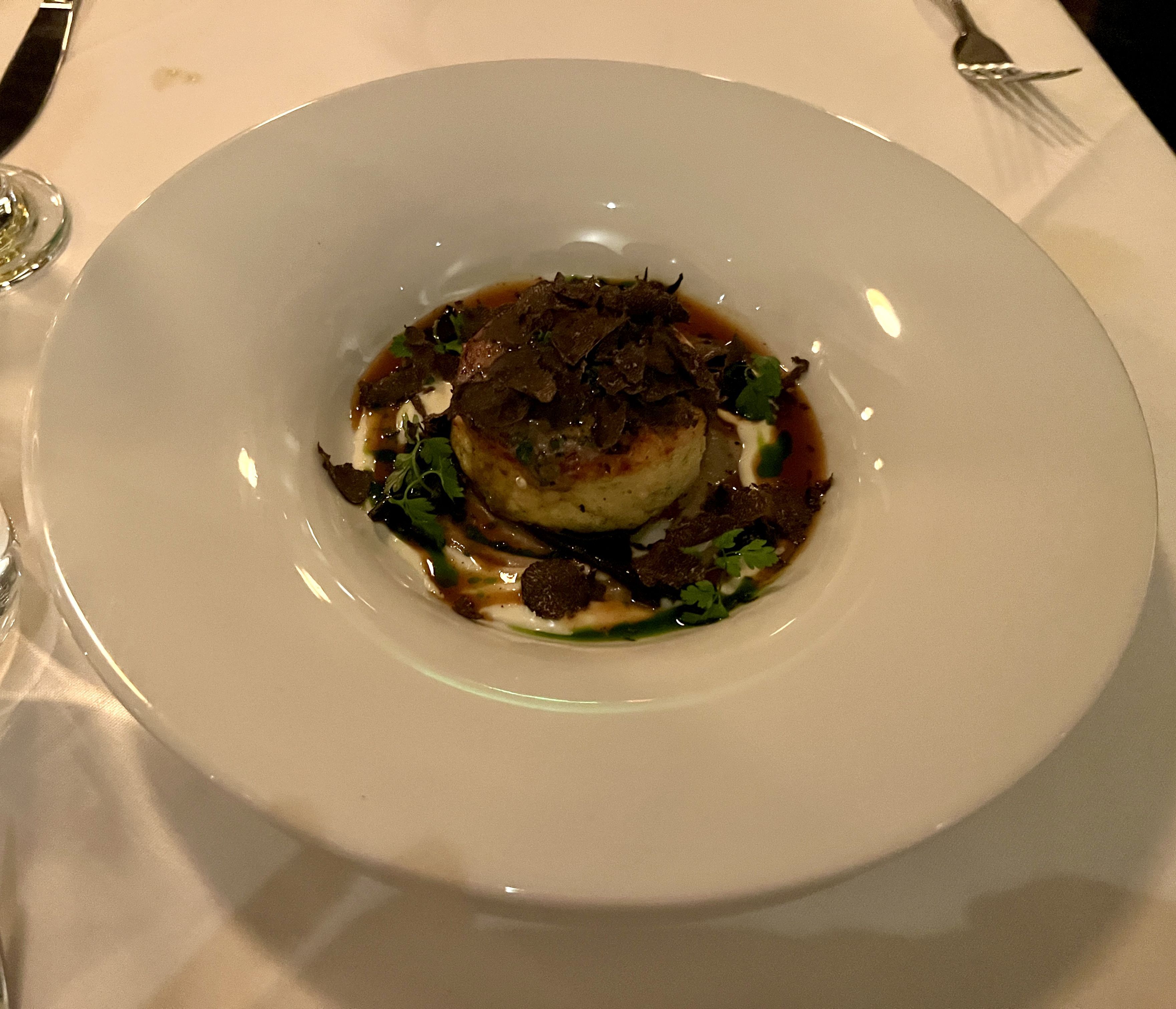 Top-down view of a plated dish in a white bowl with dark sauce, shaved black truffles, green herbs, and a creamy dollop; white tablecloth and silverware visible.