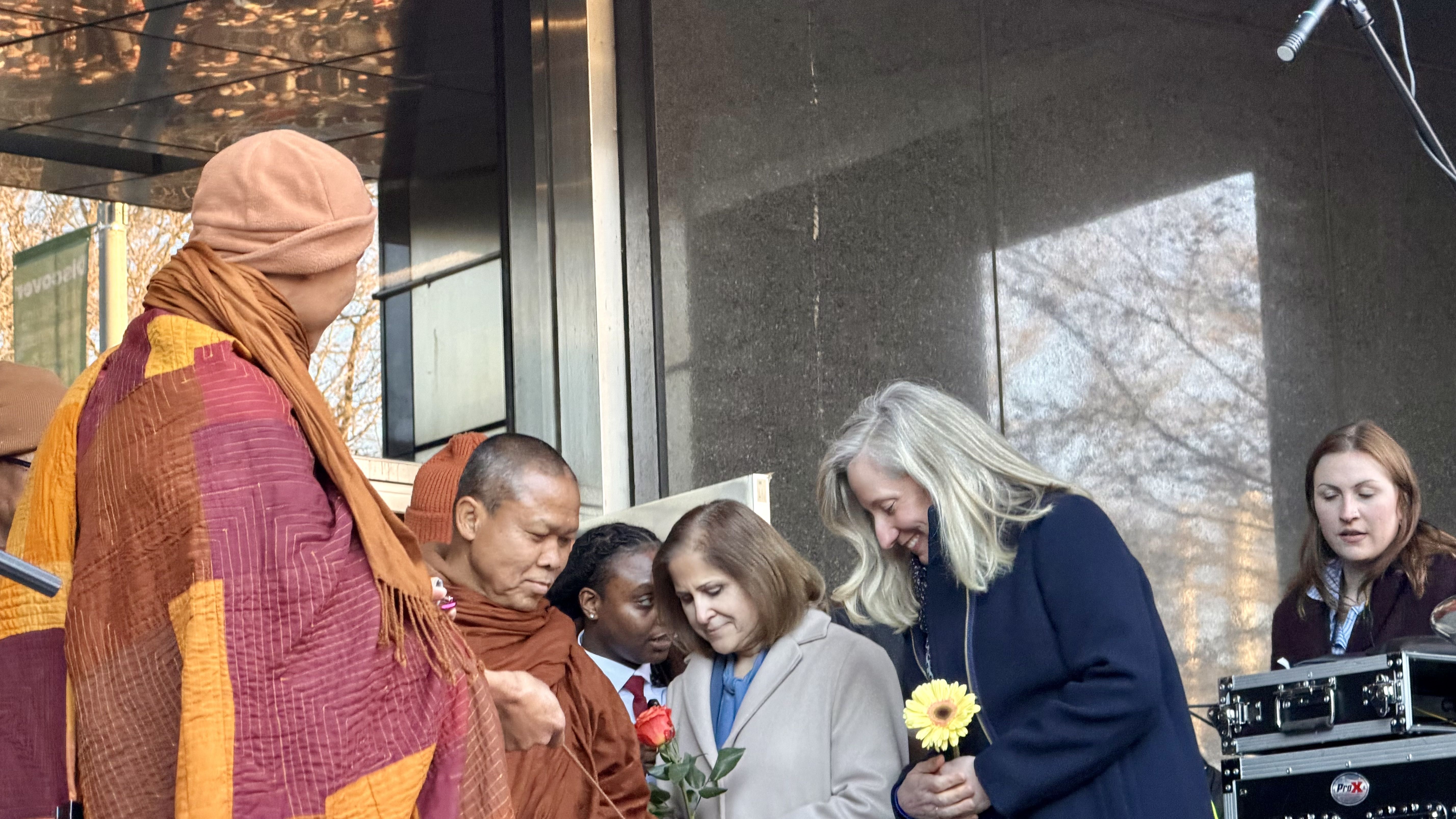 Group of people including Buddhist monks in orange robes and two women holding flowers, one red rose and one yellow daisy, gathered near a microphone and audio equipment outdoors.