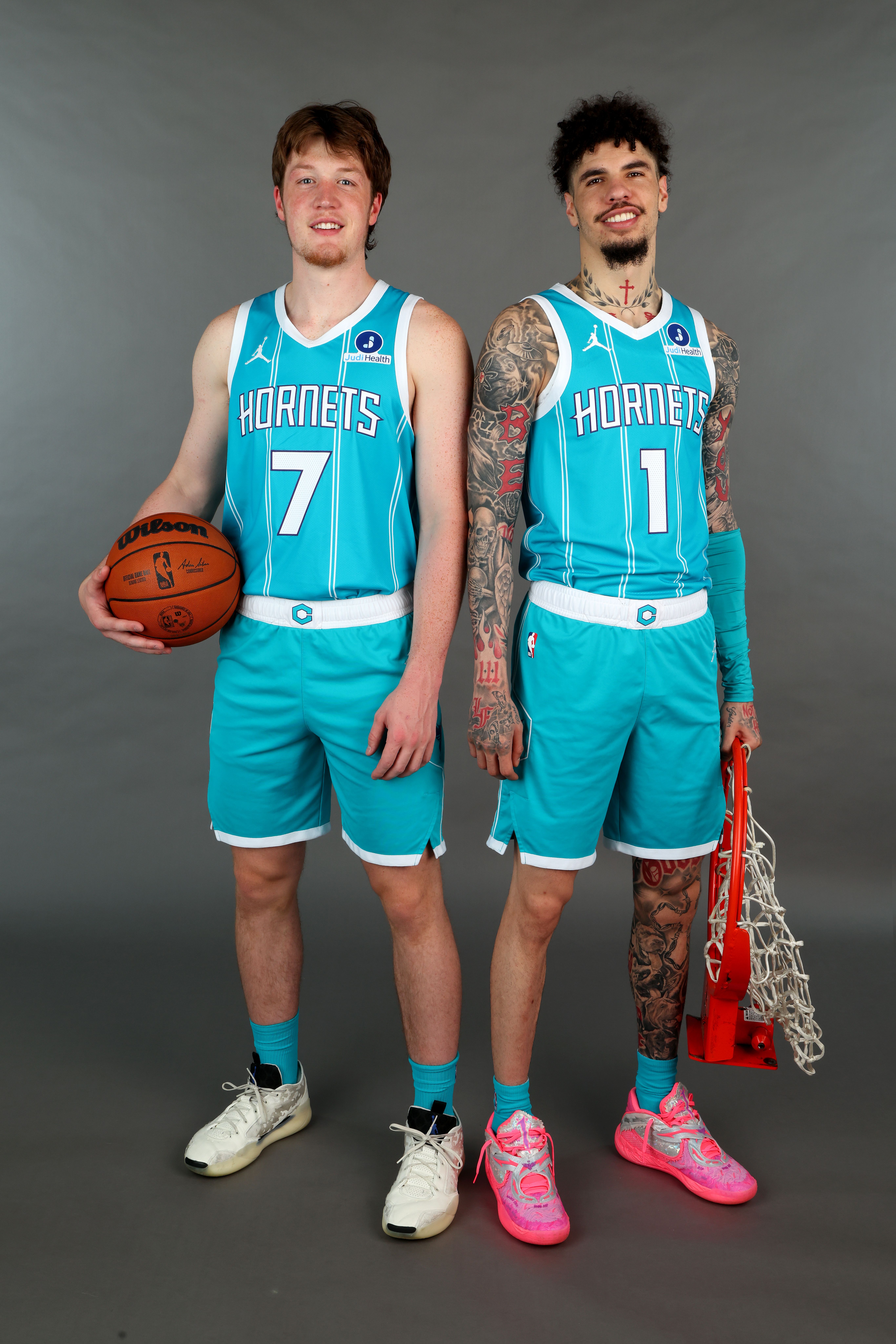 Two male basketball players in teal Hornets uniforms stand side by side against a gray background. The player on the left holds a basketball, the one on the right holds an orange net piece.