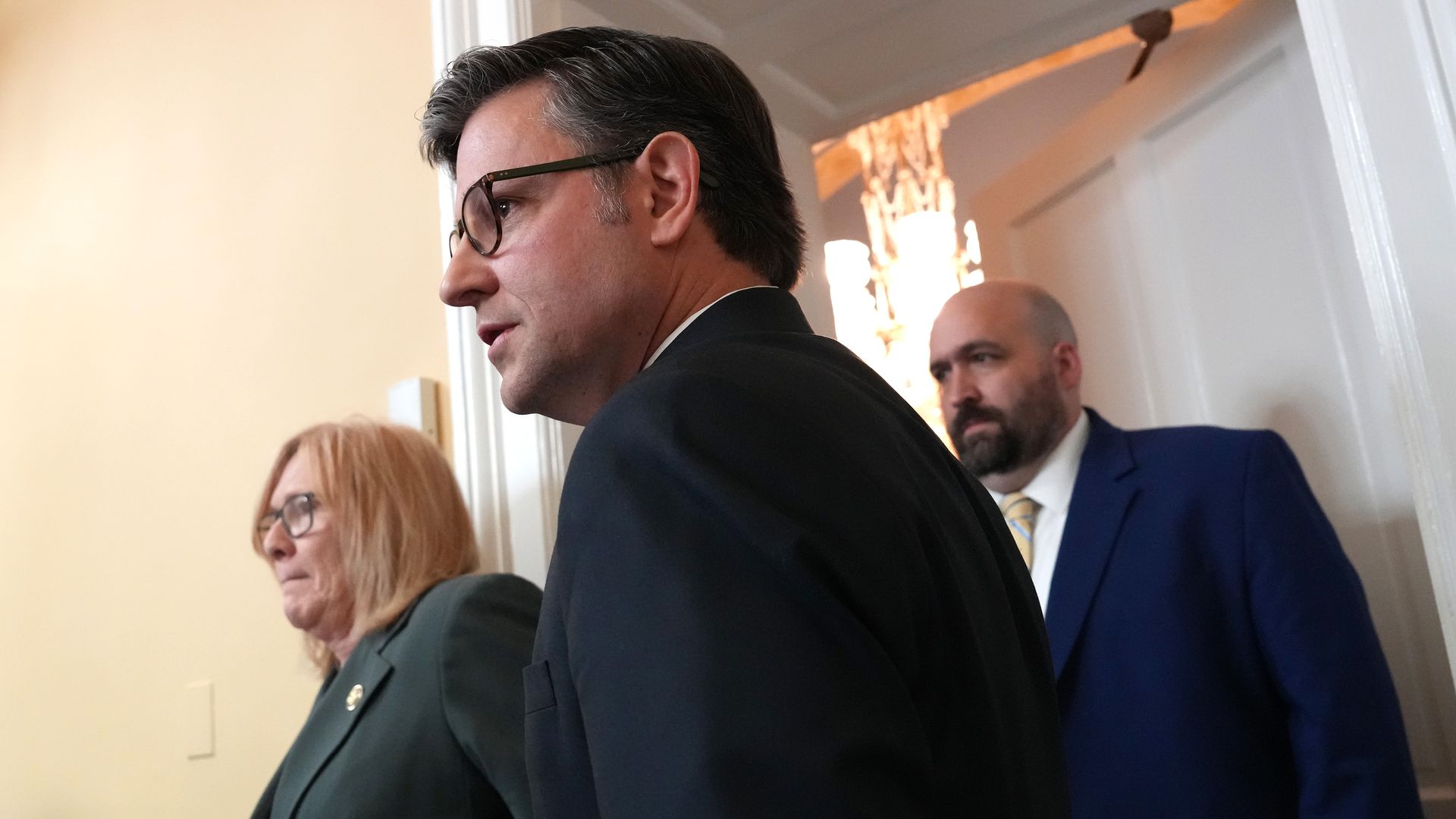 House Speaker Mike Johnson, wearing a dark suit and standing next to two people in a white room with a white doorway.