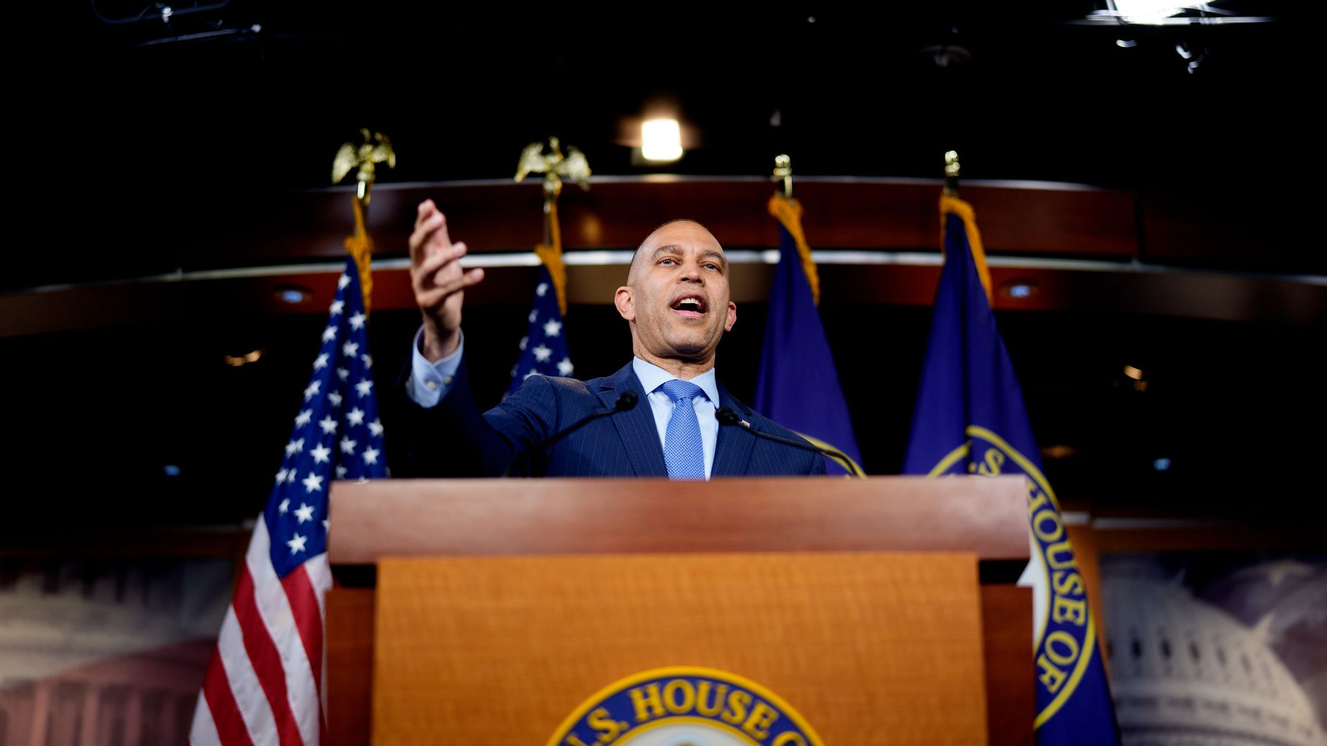House Minority Leader Hakeem Jeffries, wearing a blue suit and holding up an arm while speaking at a podium in front of a row of flags.