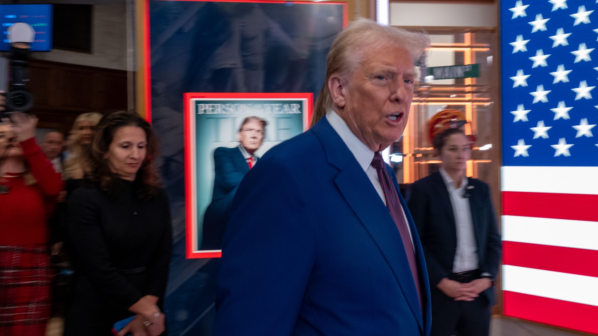 Trump enters NYSE with a group of people walking behind him. To the right, a digital American flag is partially visible. To his left, the TIME Person of the Year cover is visible. 