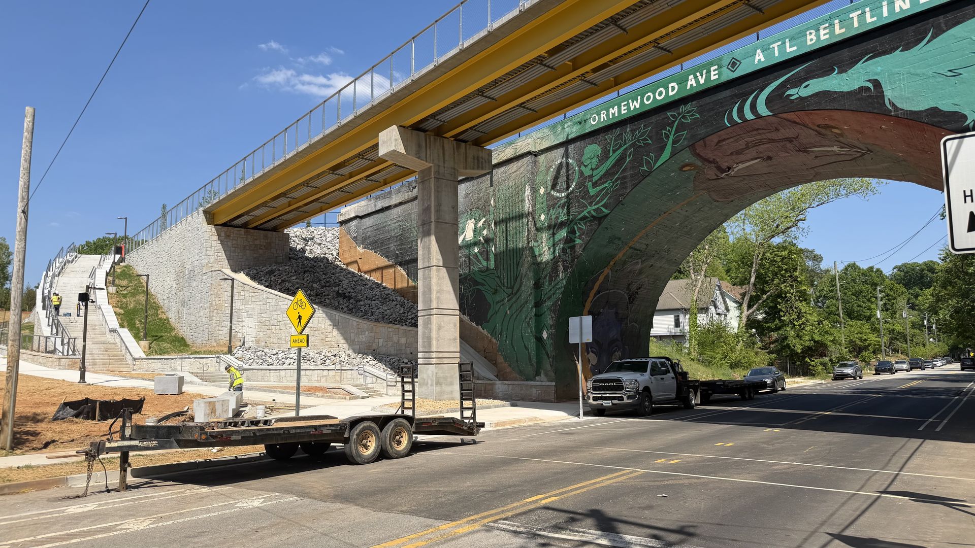 Blue sky over a street beneath a yellow steel bridge with a teal-green mural on the arch. Construction workers in bright vests, a flatbed trailer, stone wall, stairs, and parked cars line the road.