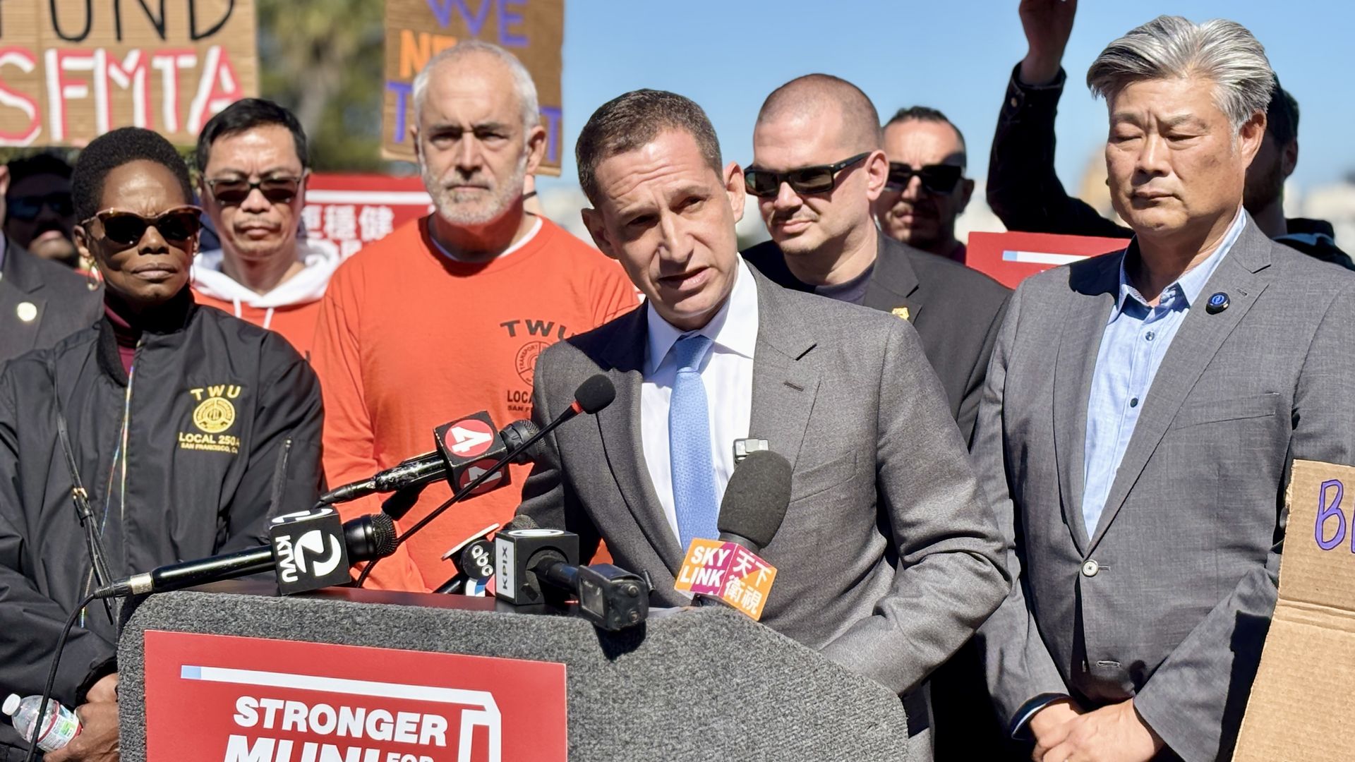 Man in gray suit and blue tie speaks at podium with microphones, surrounded by diverse group of people wearing TWU Local 250-A gear and holding signs outdoors on sunny day.