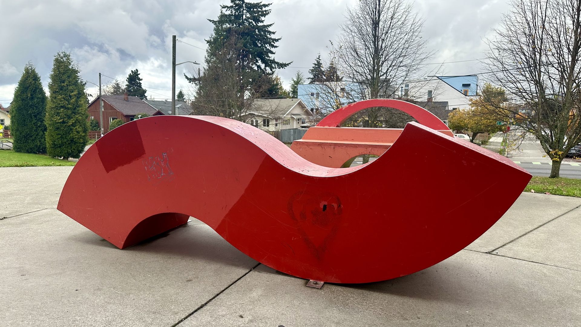 A large red wavy sculpture is on paving stones with bare trees and evergreens in the background. 
