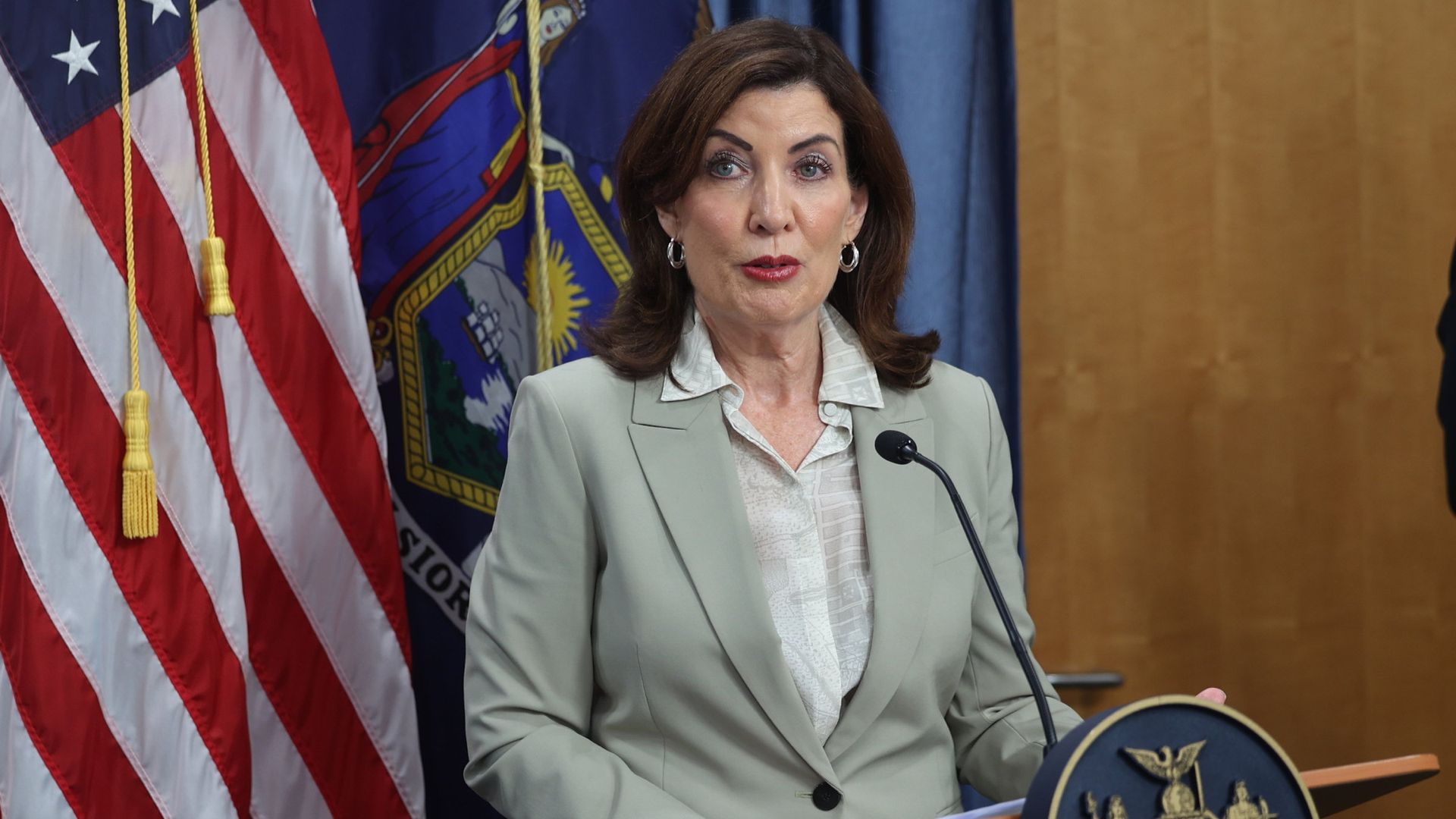 New York governor Kathy Hochul  in a light gray suit speaking at a podium with New York state seal, American flag, and New York state flag in the background.