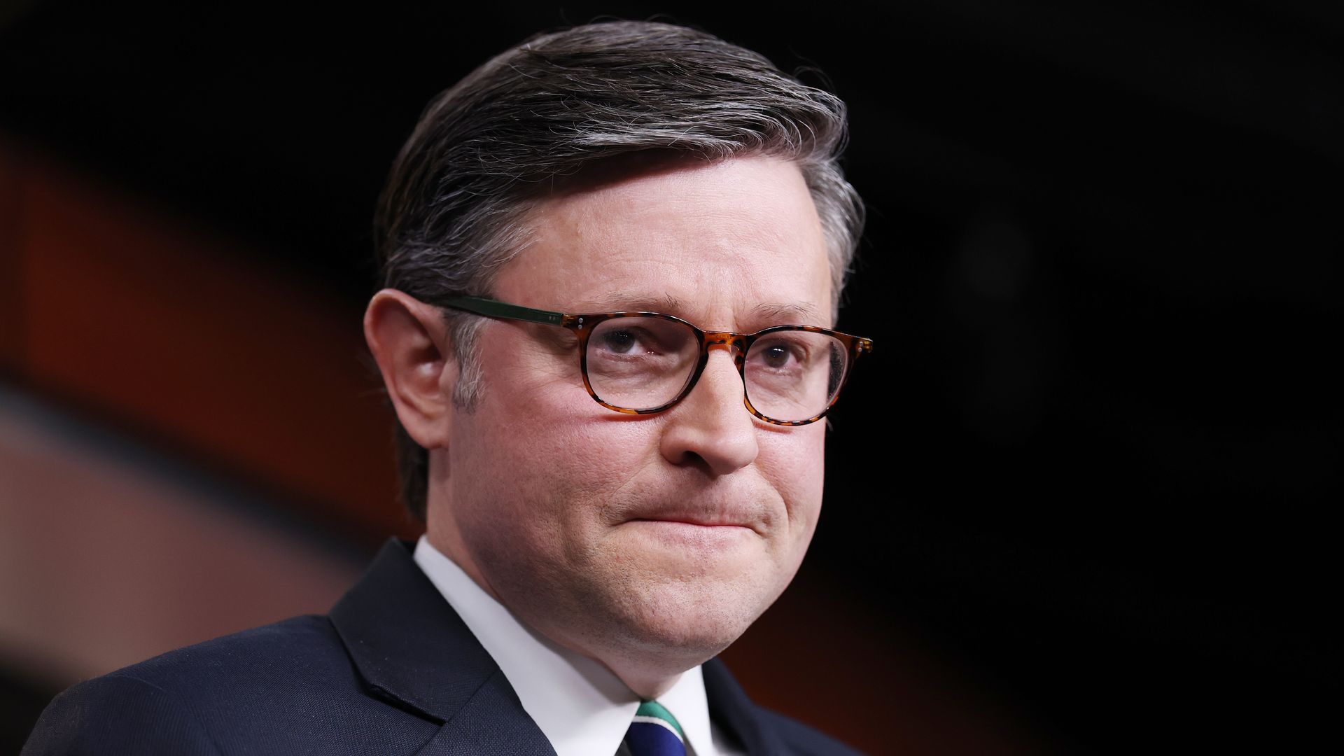 Portrait of a man with gray hair and tortoiseshell glasses, in a dark suit, white shirt, and green striped tie, with an American flag lapel pin, speaking at a podium.