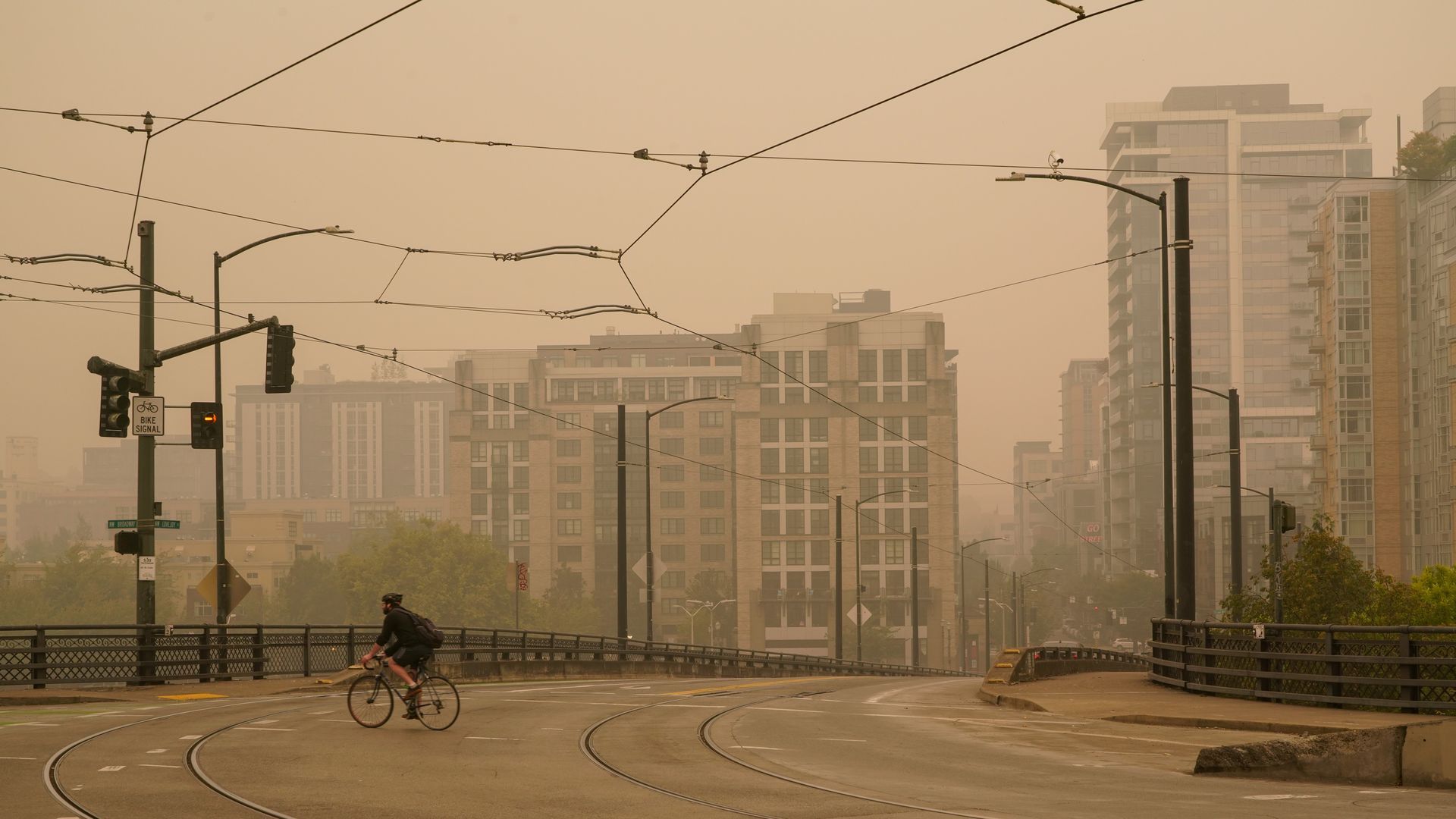 A person rides a bike through an intersection with a background of thick, orange-like smoke from a nearby wildfire hangs in the background.